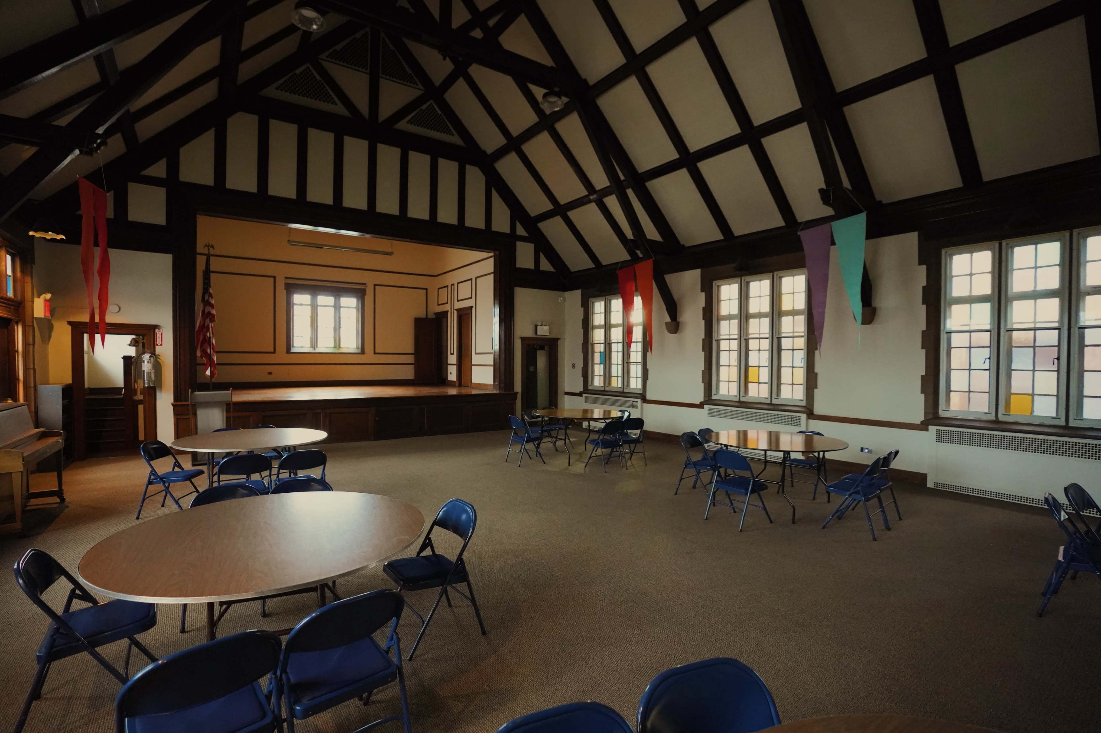 The image shows an empty hall with several round tables and blue chairs arranged around a stage at the front.