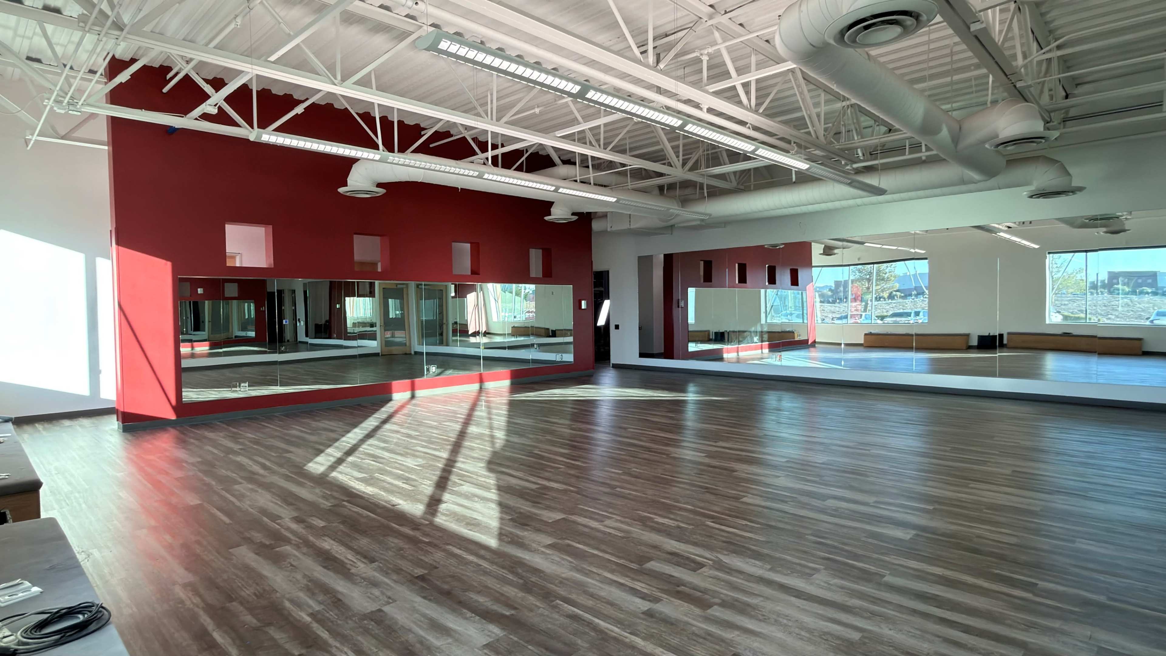 The image shows a spacious room with hardwood floors, large mirrors on one wall, and red accent walls, illuminated by natural light from large windows.