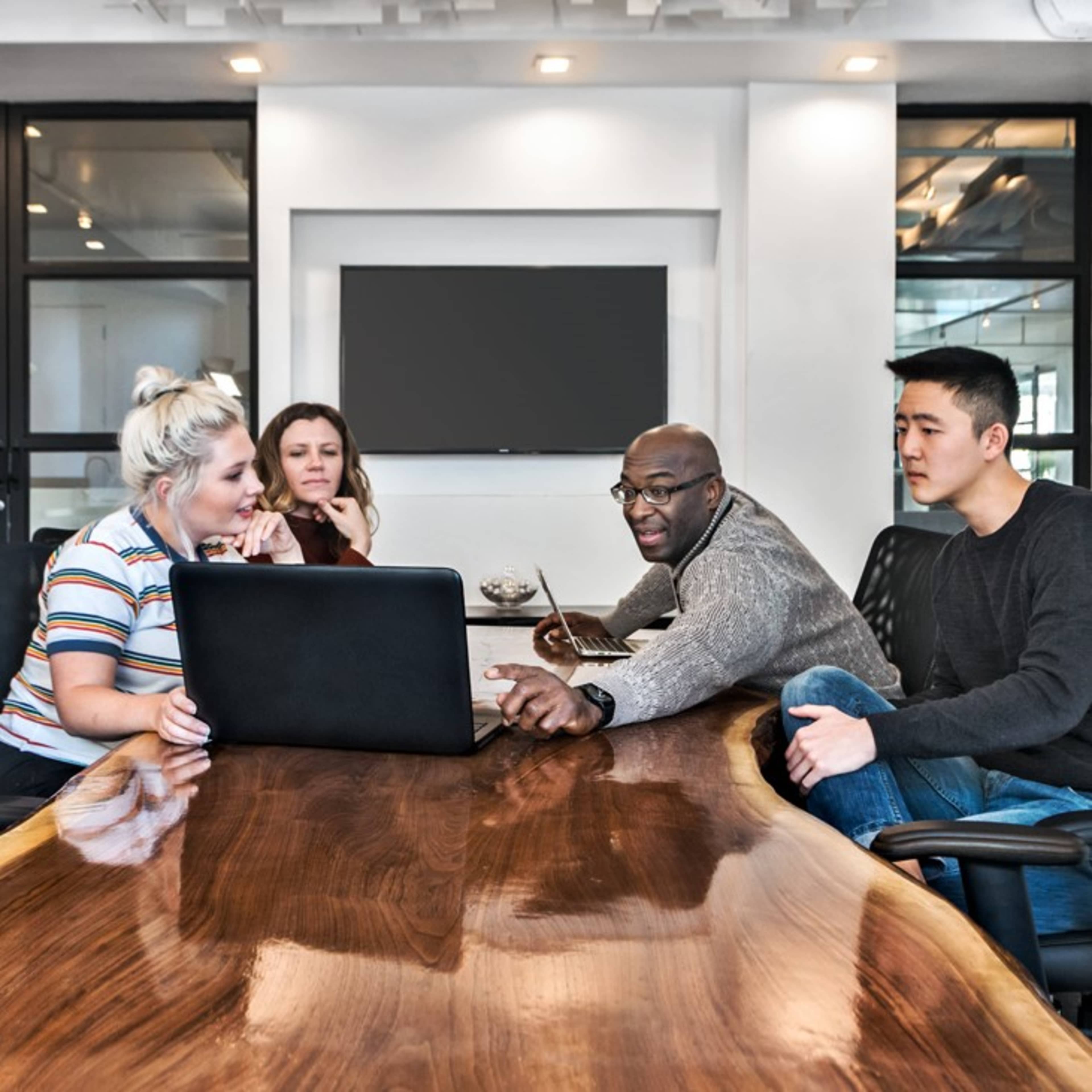 A group of four people sitting around a large wooden table, looking at a laptop in a modern office setting.