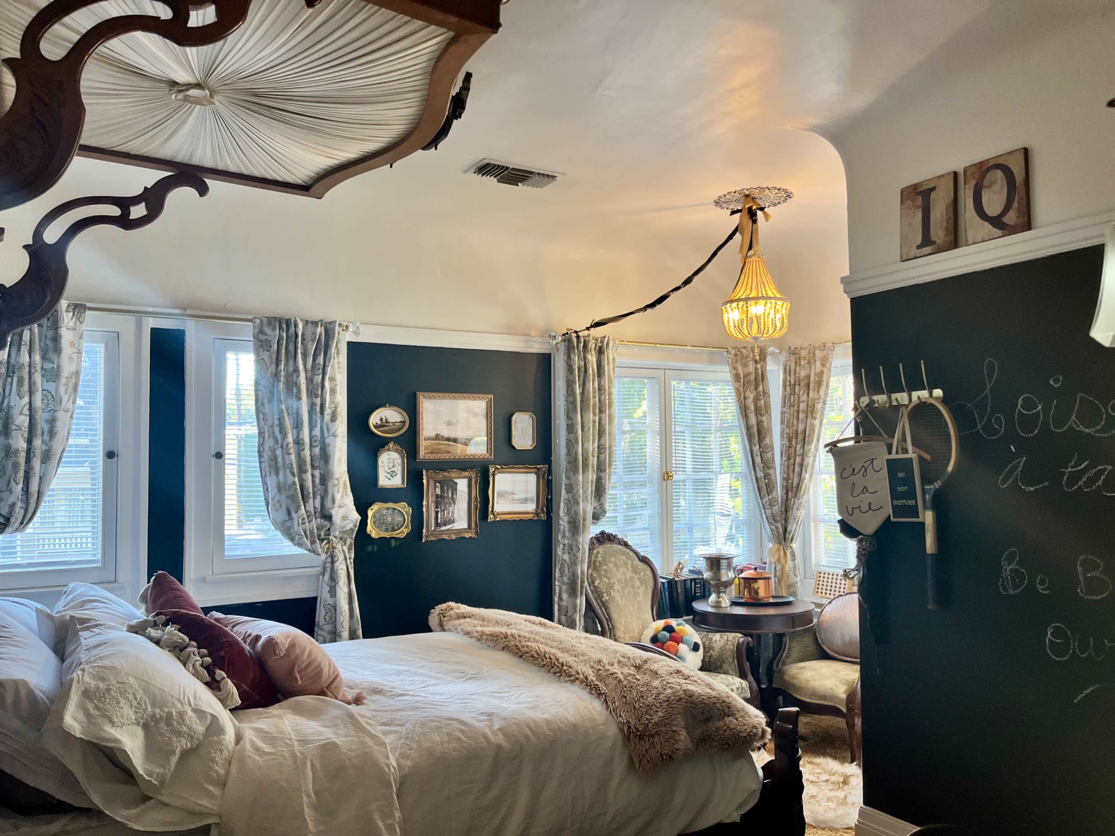 A cozy bedroom with a white bed, decorative wall hangings, a chandelier, and a table set near large windows.