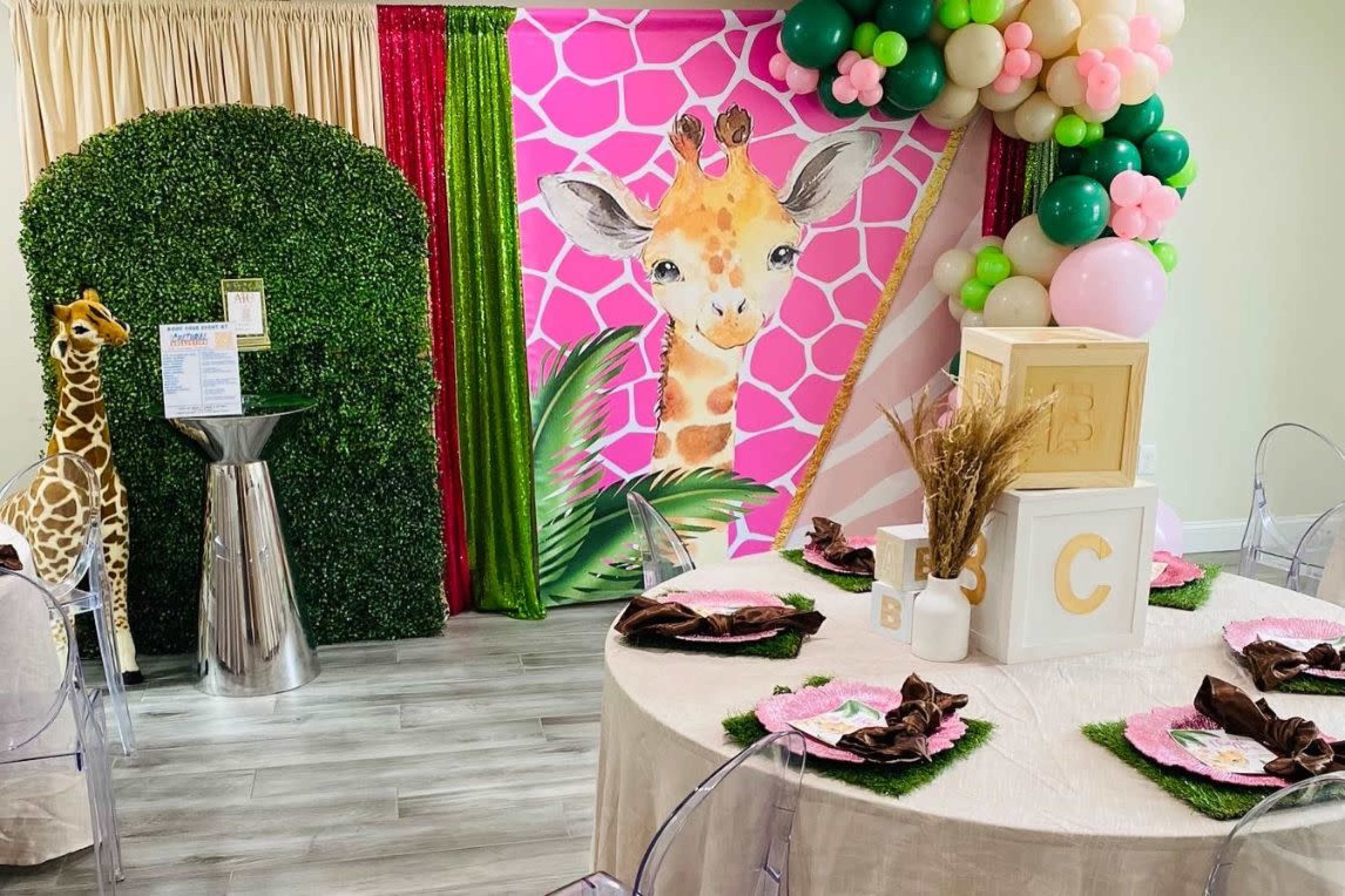 A decorated event space featuring a large pink giraffe mural, a round table with patterned tableware, and a colorful balloon arch.