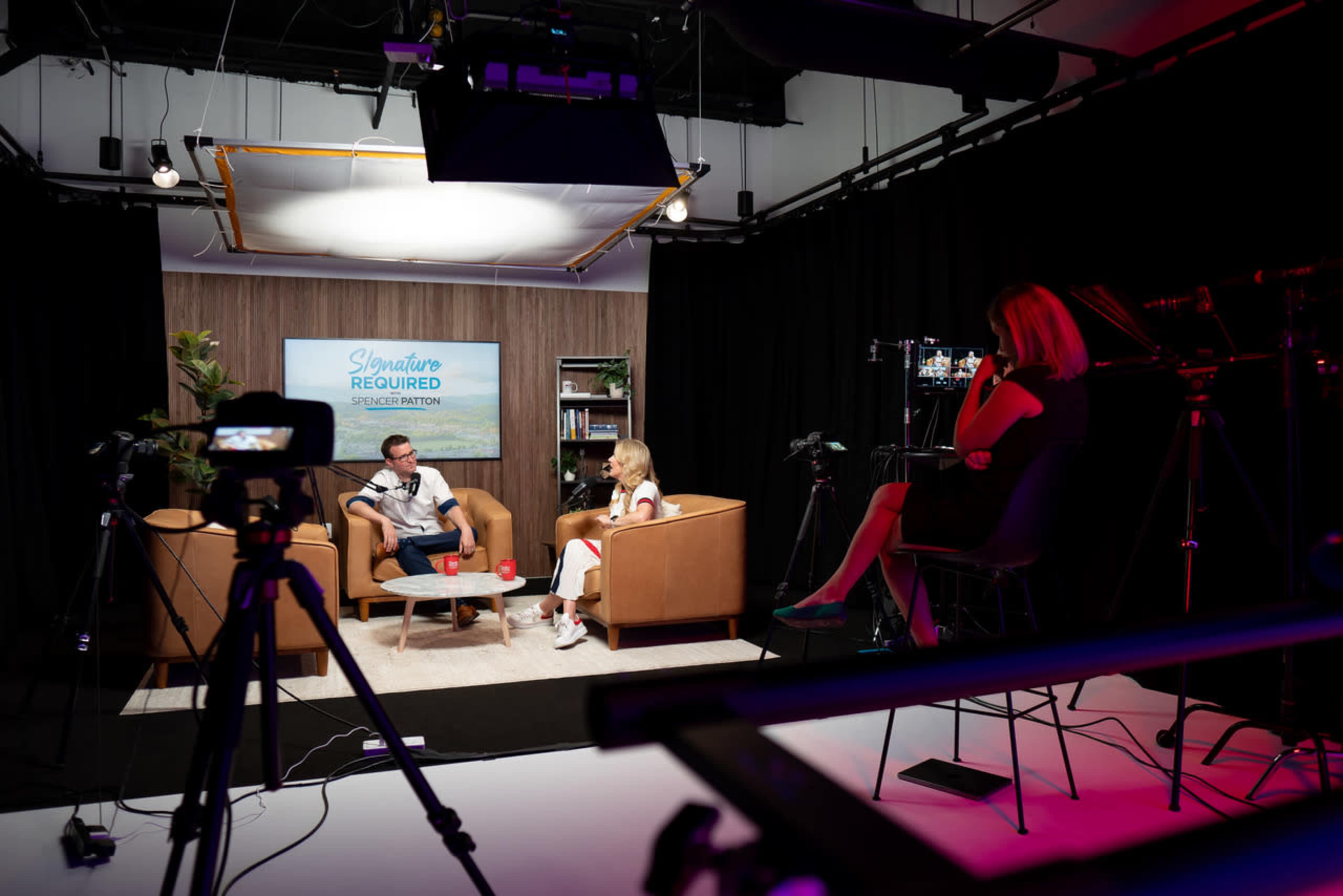 A studio setup features two people seated on couches engaged in a conversation, while cameras and lighting equipment are arranged around the scene.