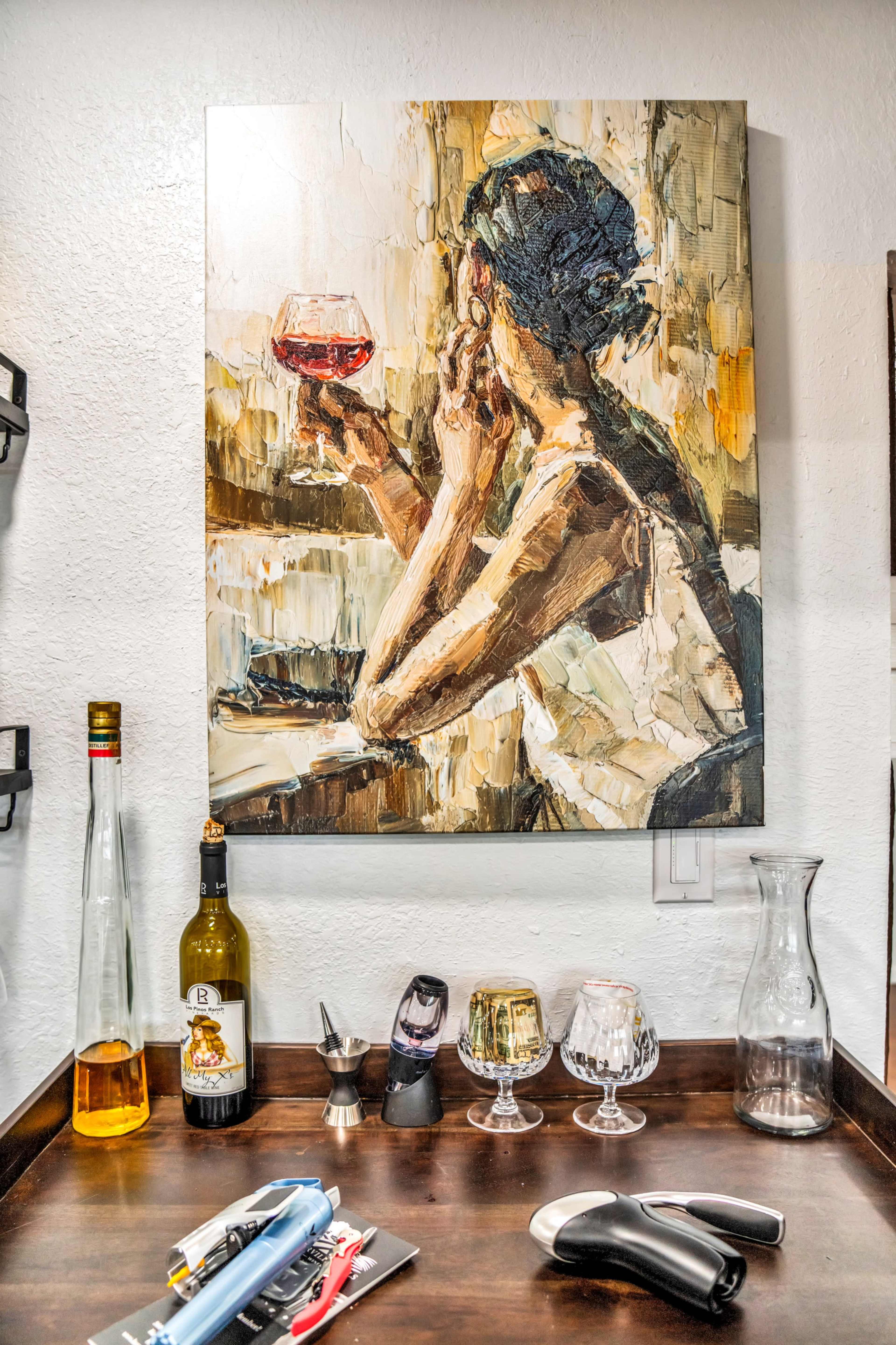 A wooden counter displaying various glassware and bottles, with a large painting of a woman holding a glass of red wine hung on the wall above.