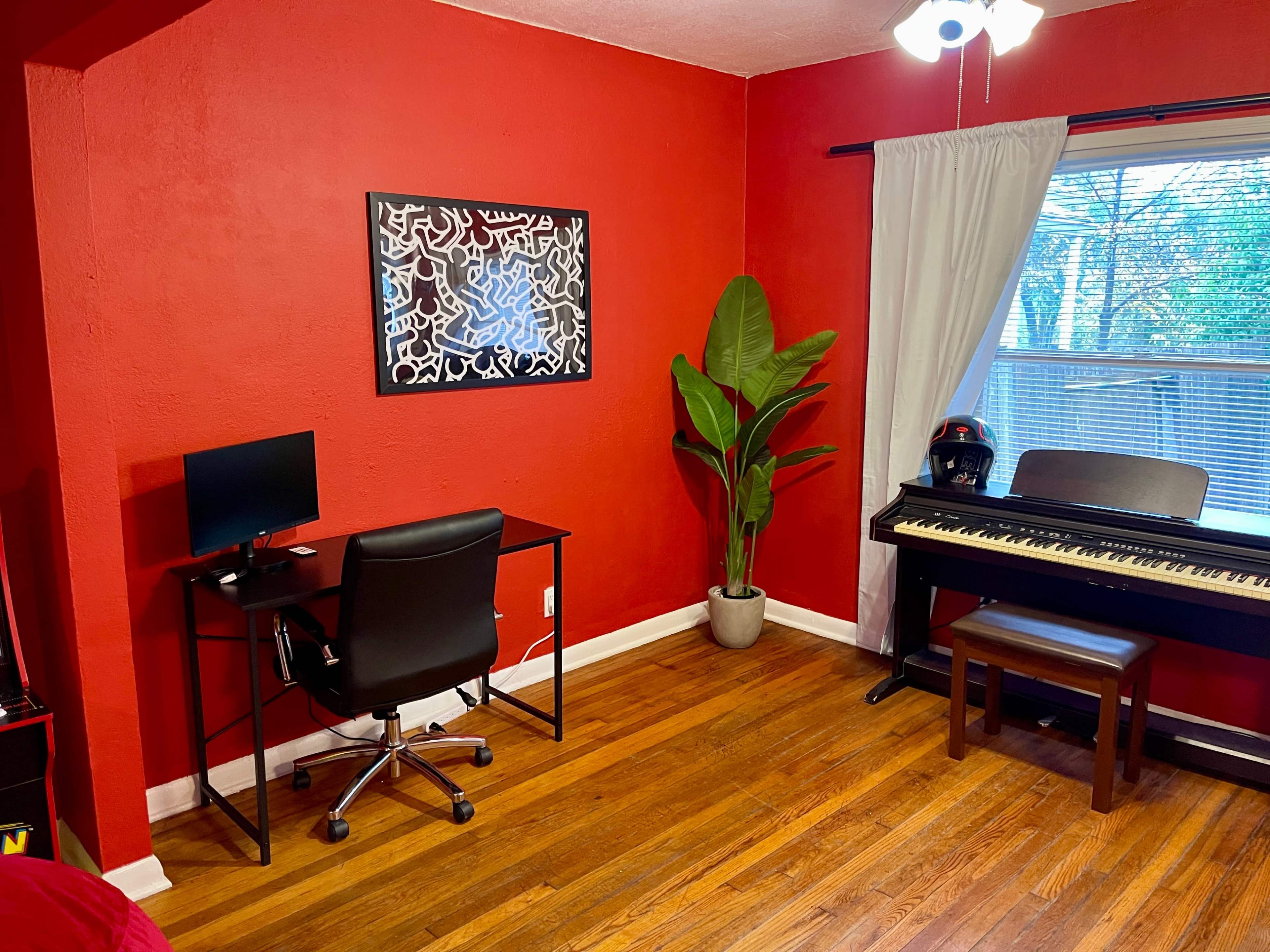 The image shows a room with red walls featuring a desk with a computer monitor, a keyboard piano, a chair, and a large plant near a window.