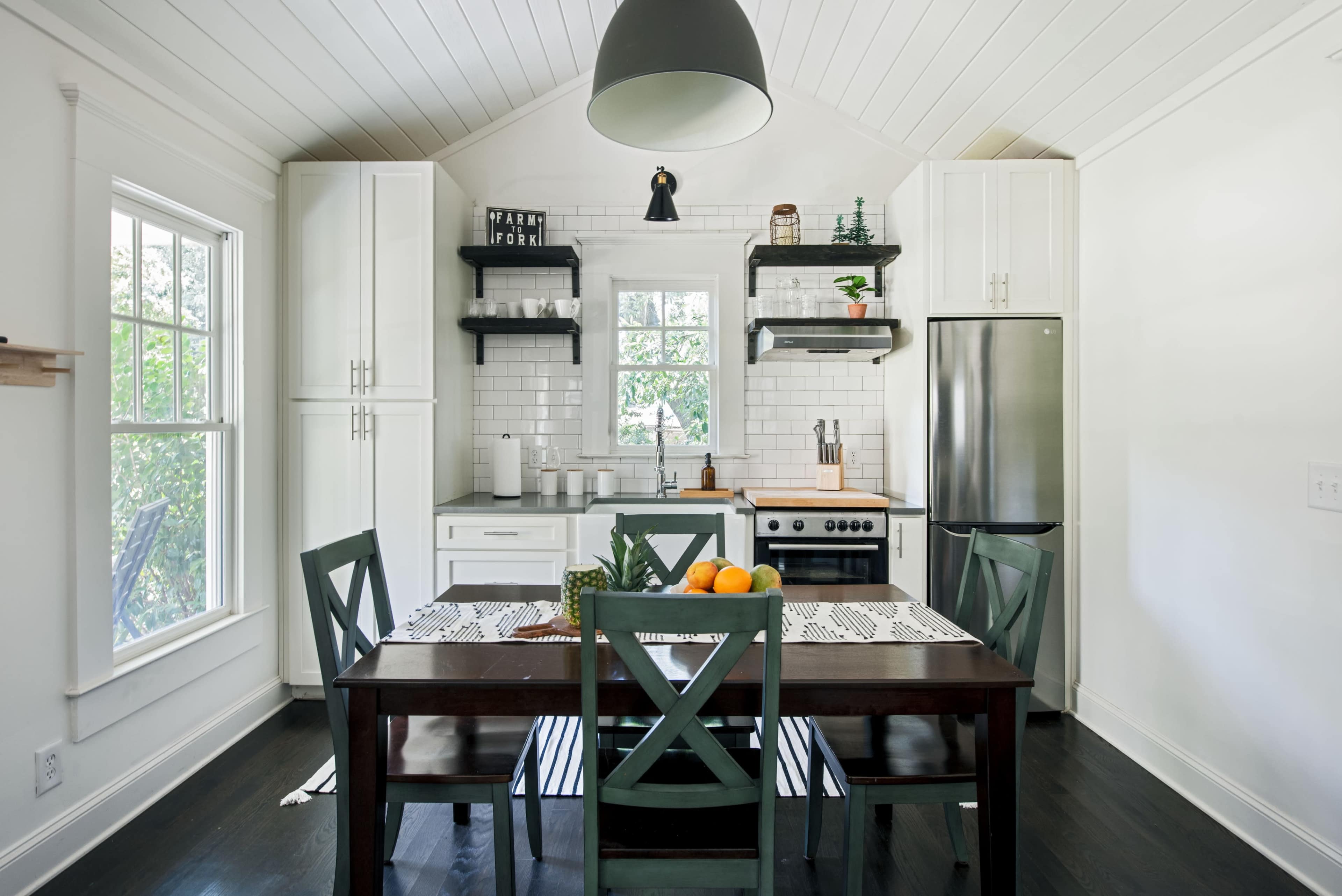 A modern kitchen features a central dining table with green chairs, white cabinets, and stainless steel appliances, illuminated by a hanging light fixture.