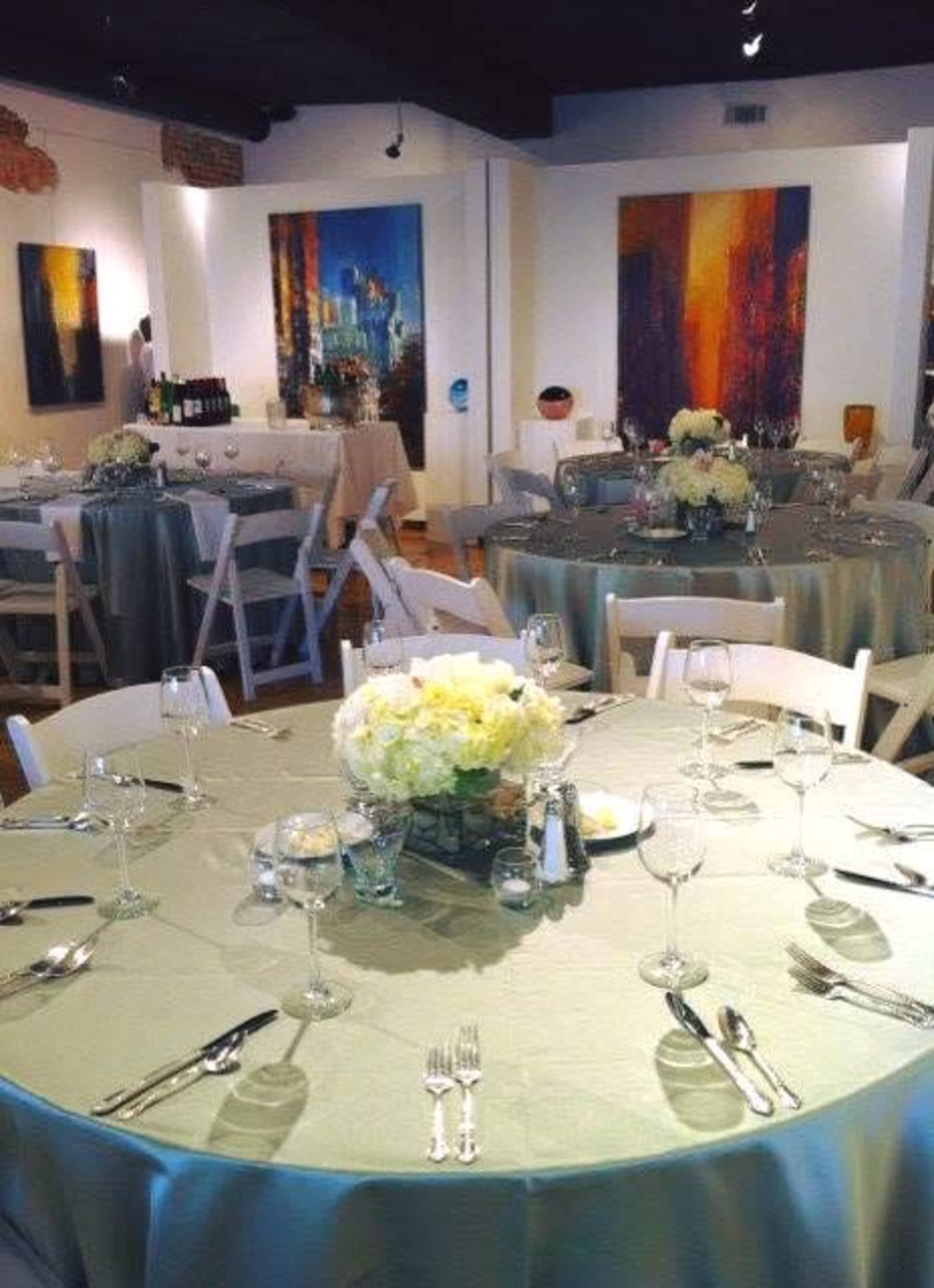 The image shows a banquet hall set up for an event, featuring round tables with light-colored tablecloths, centerpieces of flowers, and elegant place settings.