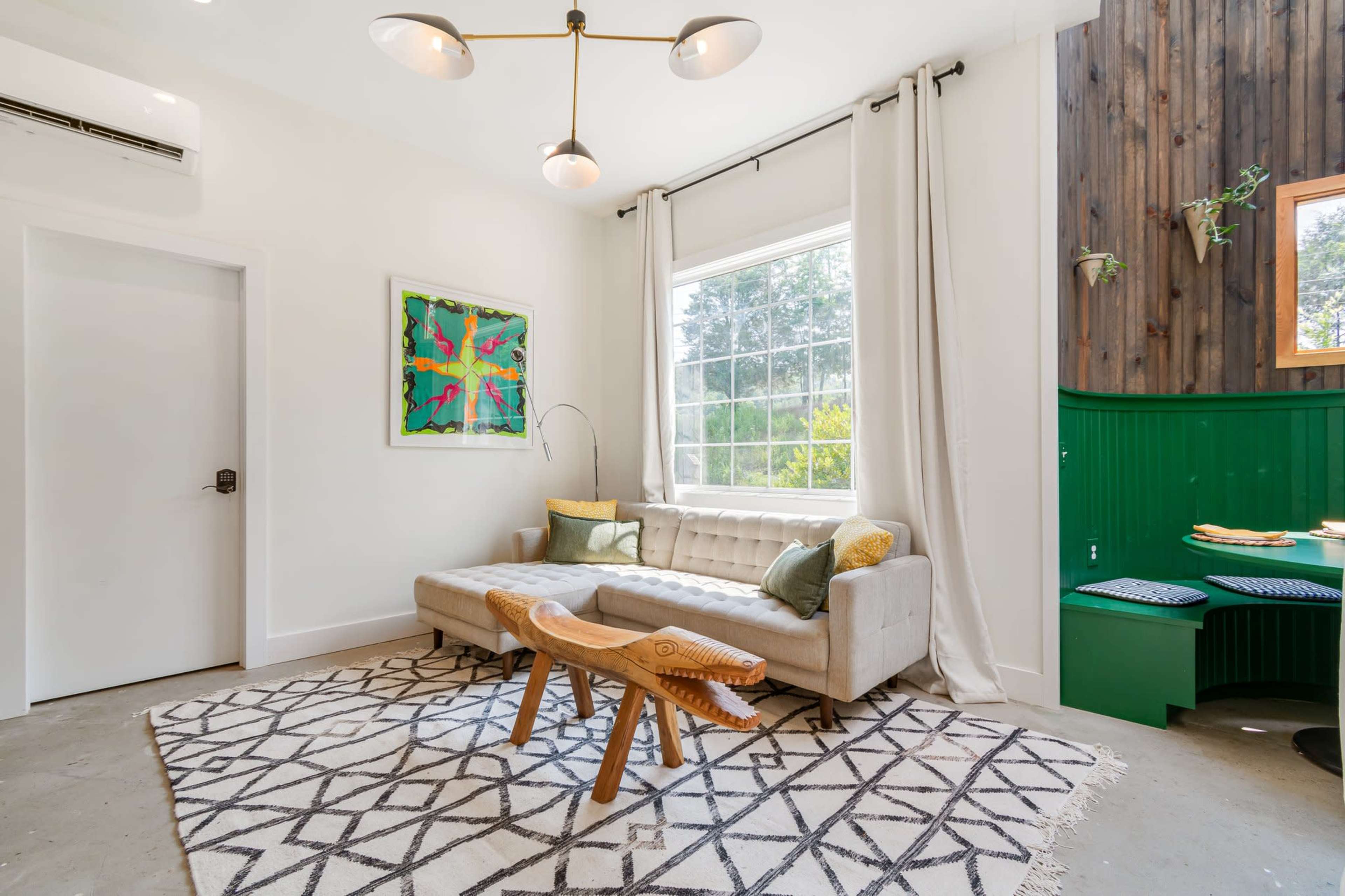 The room features a light-colored sofa with a unique wooden coffee table, large windows, and a green accent wall.