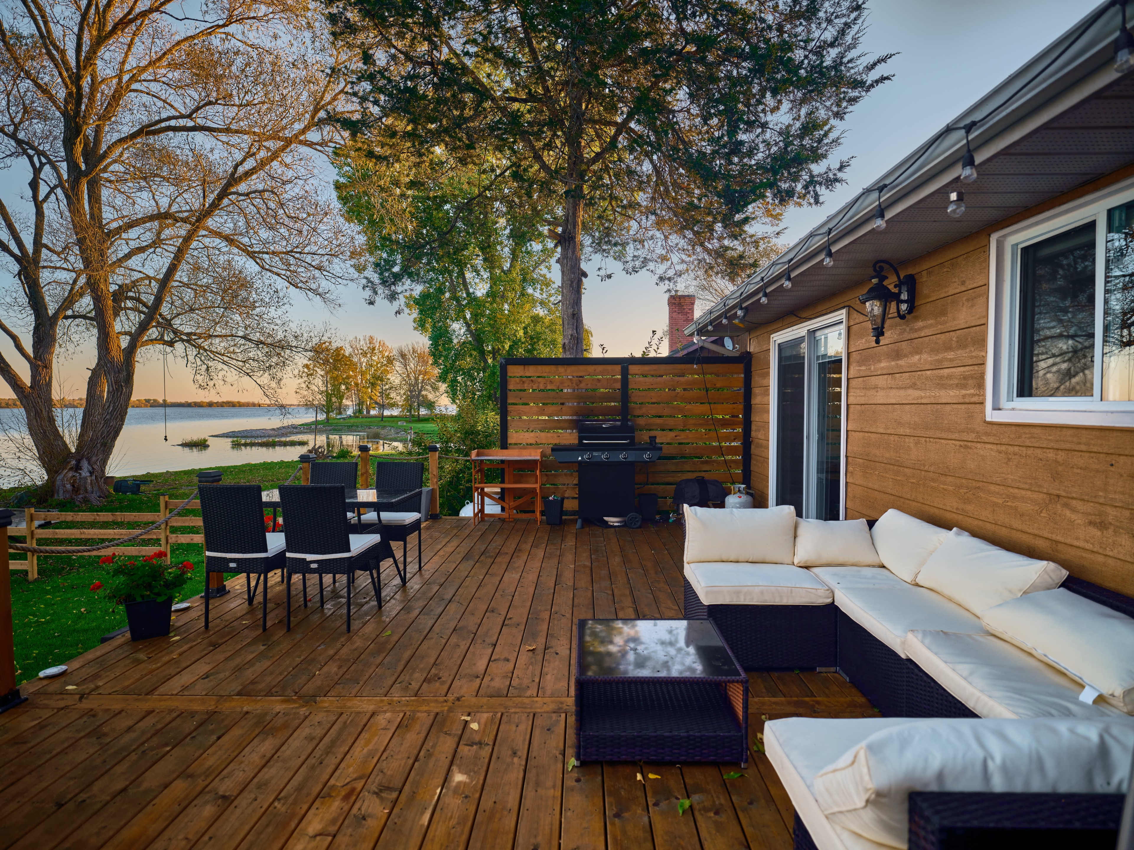 A wooden deck with outdoor seating furniture overlooking a calm body of water and trees in the background.