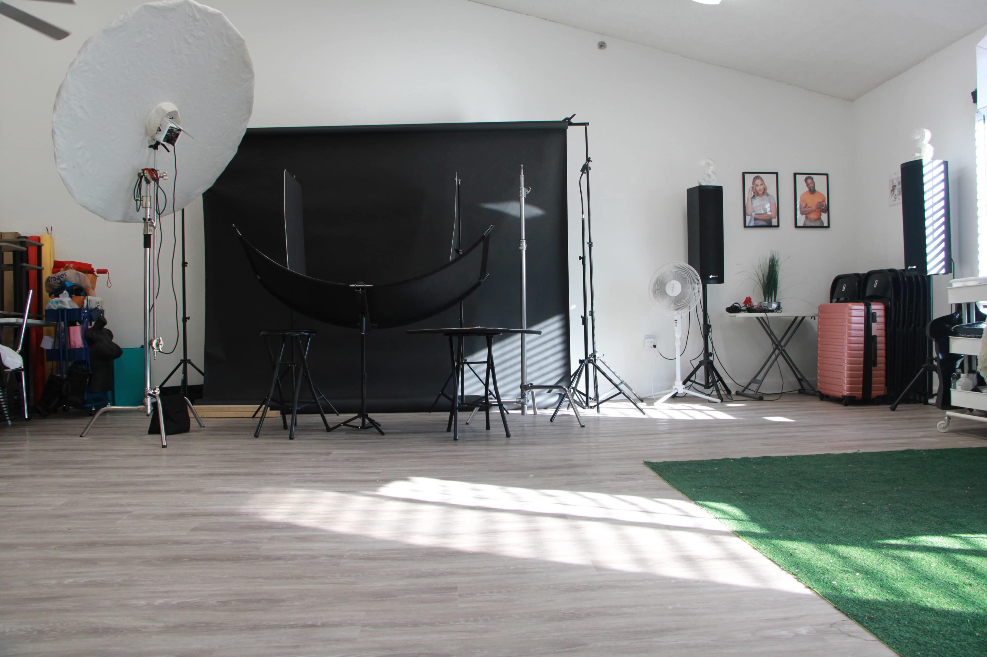 The image shows a photography studio with a black backdrop, lighting equipment, two chairs, and artificial grass on the floor.