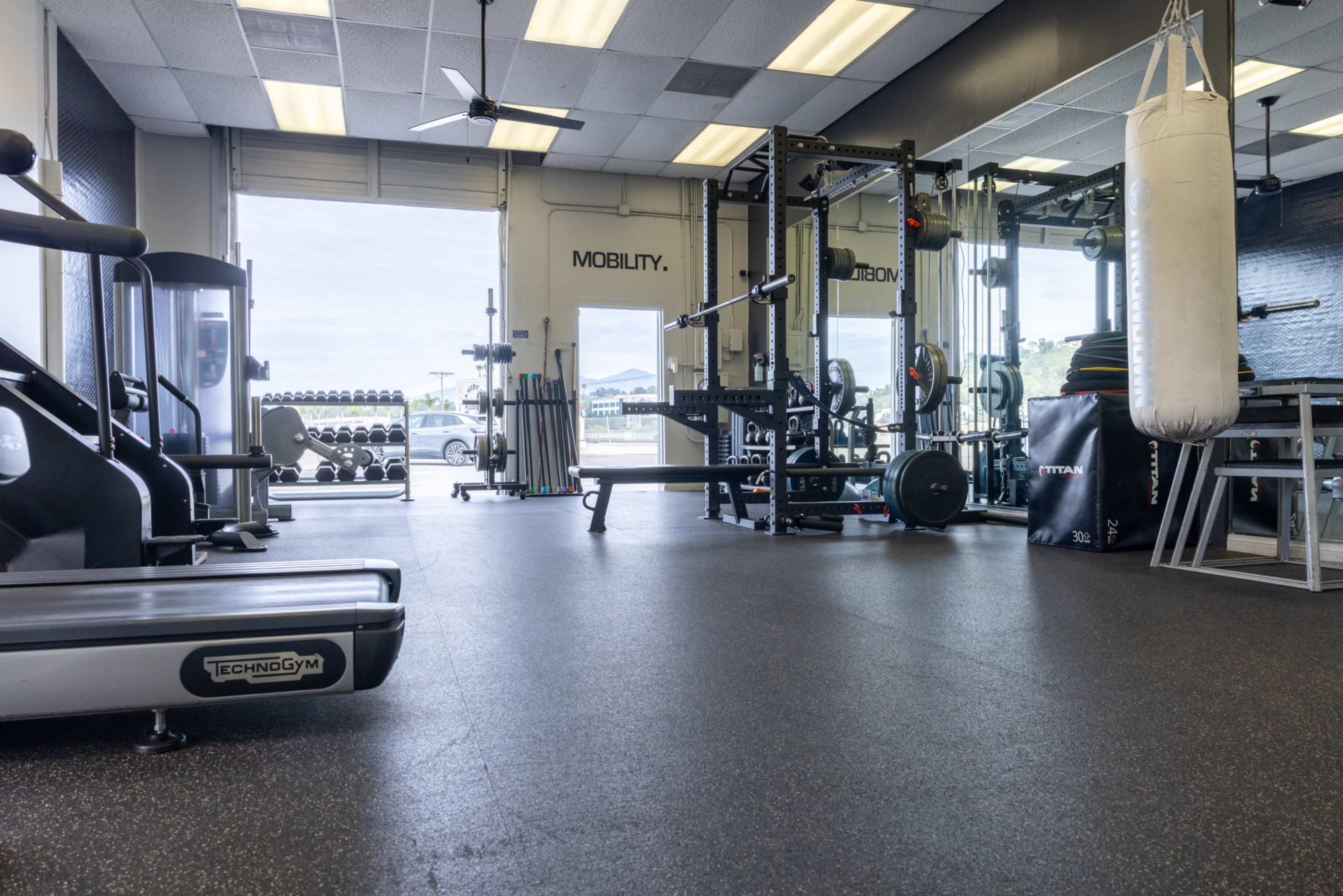 The image shows a gym interior equipped with various exercise machines, weights, and open space towards large windows.