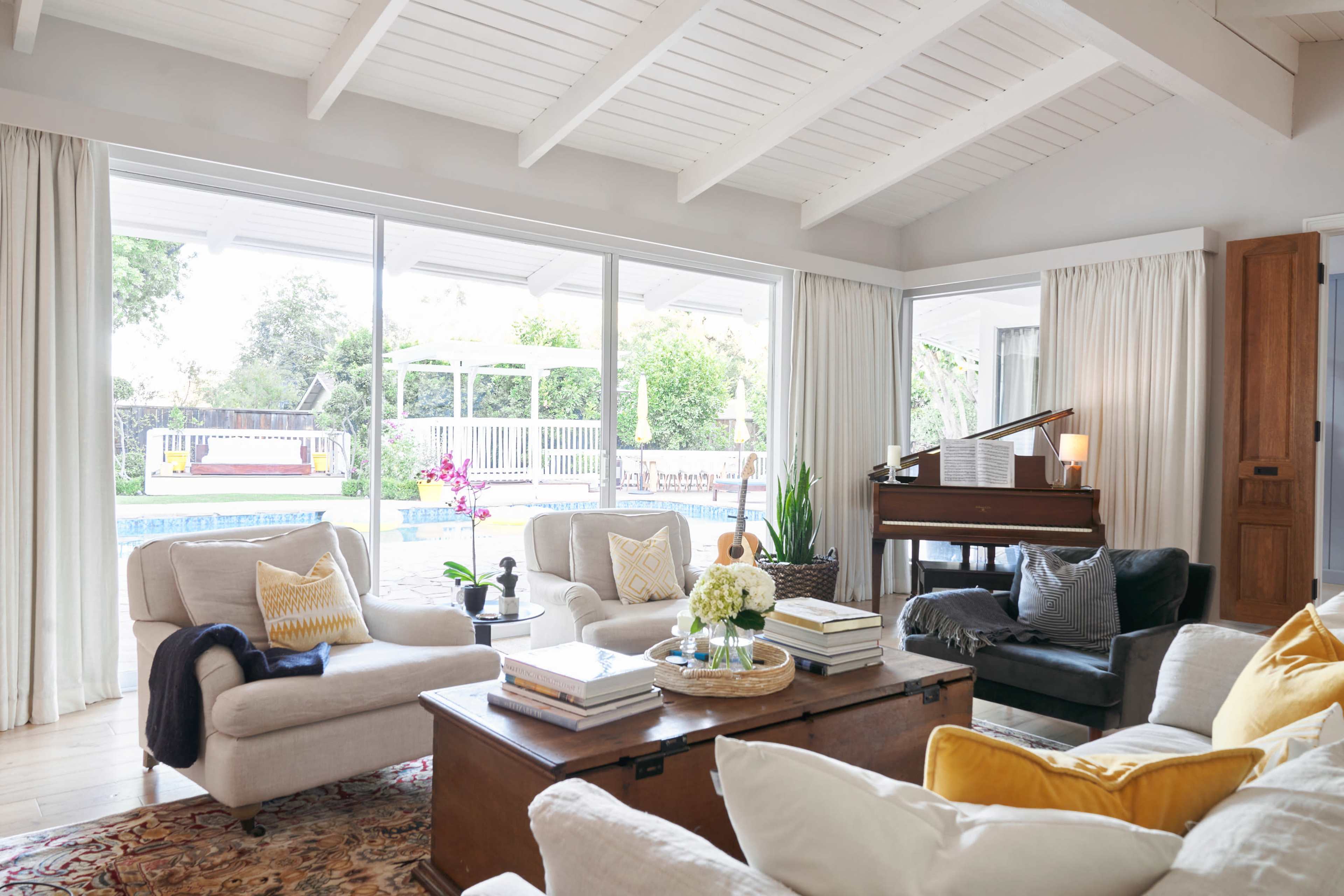 A bright living room features a piano, comfortable seating, and large windows overlooking a backyard with a pool.