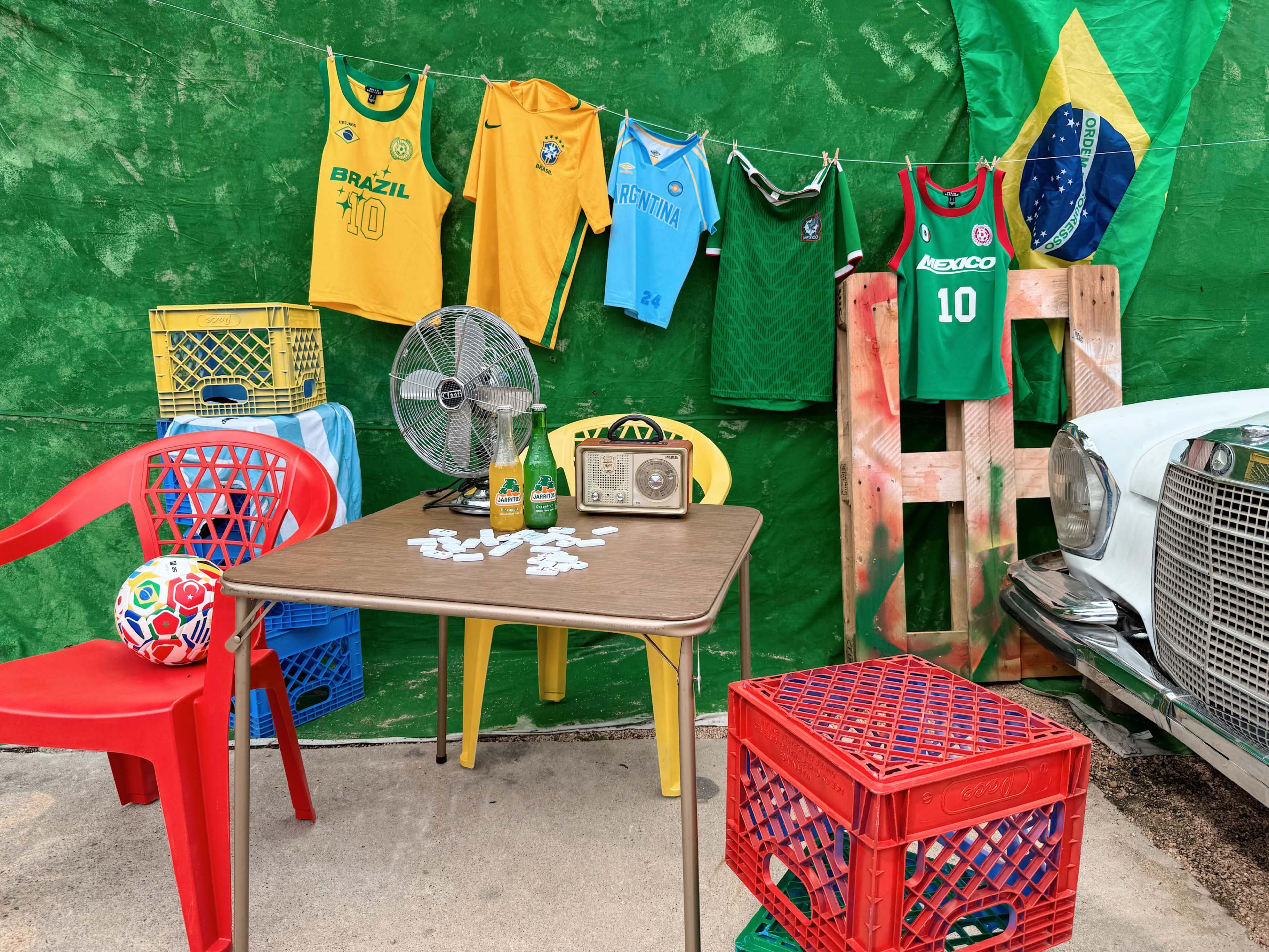 The image features a vibrant display of colorful jerseys from Brazil, Argentina, and Mexico hanging on a green background, with a table, chairs, a fan, and a vintage car in the scene.