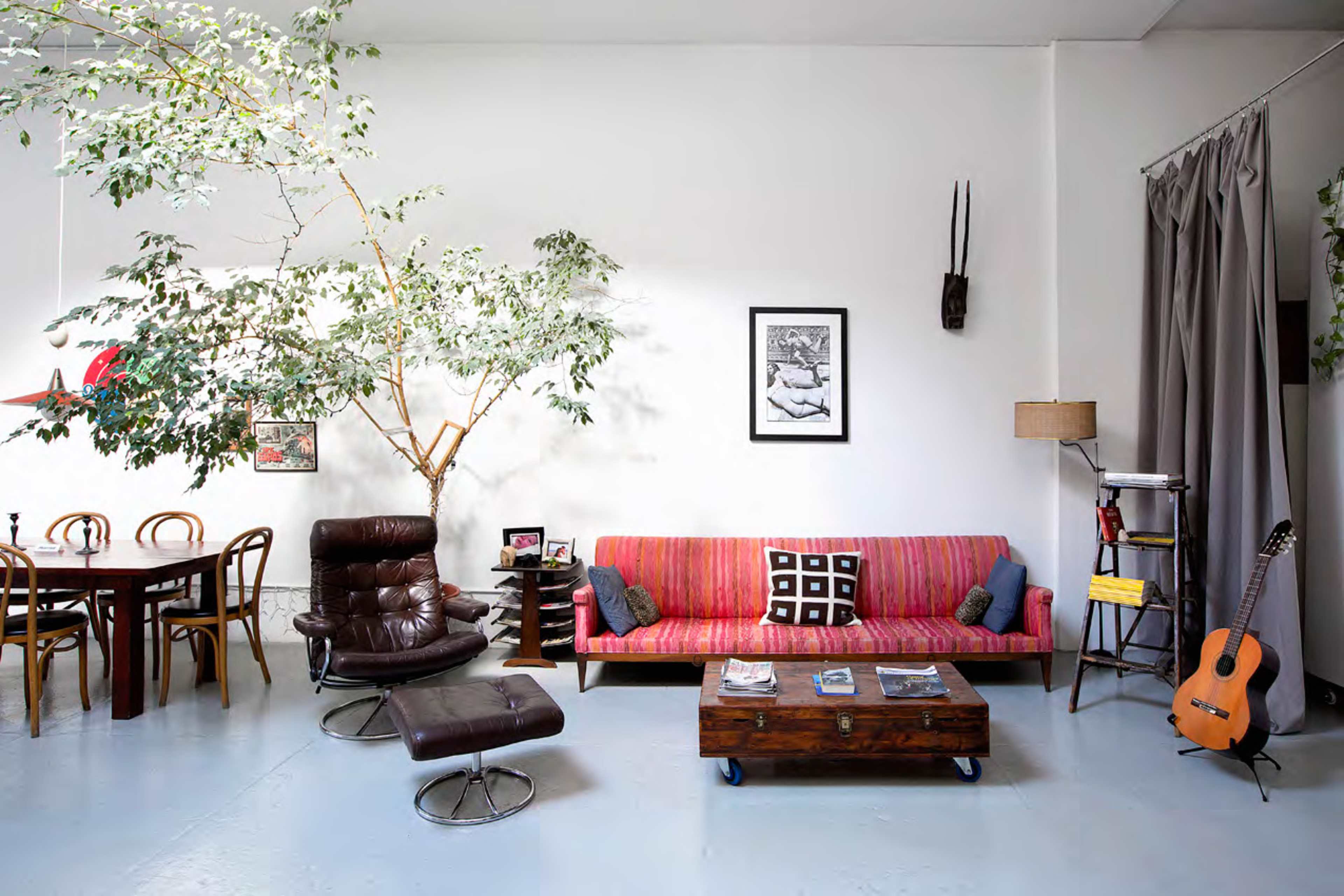 A modern living space featuring a pink-striped sofa, a wooden coffee table with magazines, a guitar, a black armchair, a dining table, and a large potted plant.