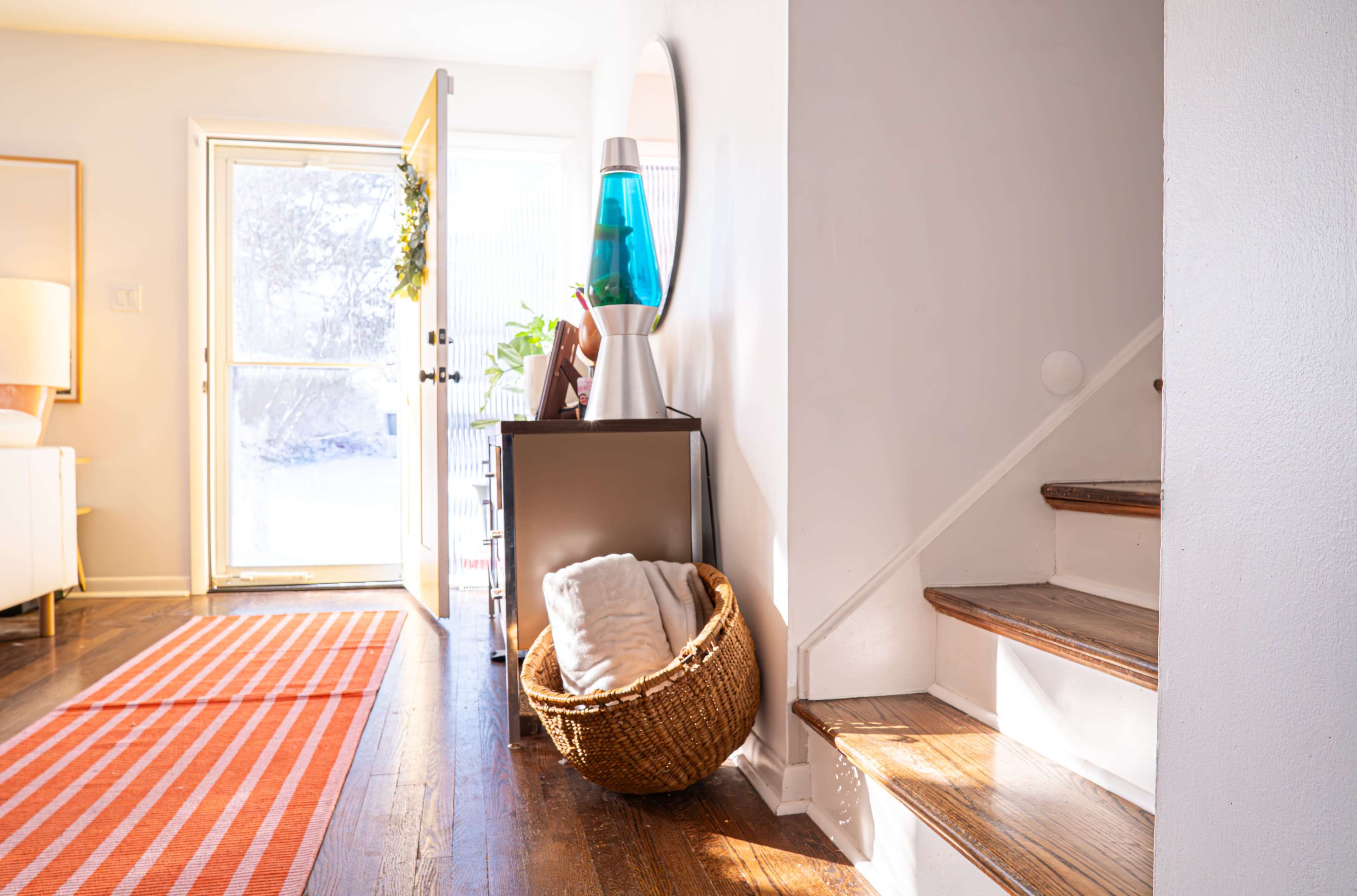The image shows a bright entryway featuring a staircase, a basket on the floor, a lamp, and a door open to the outside.