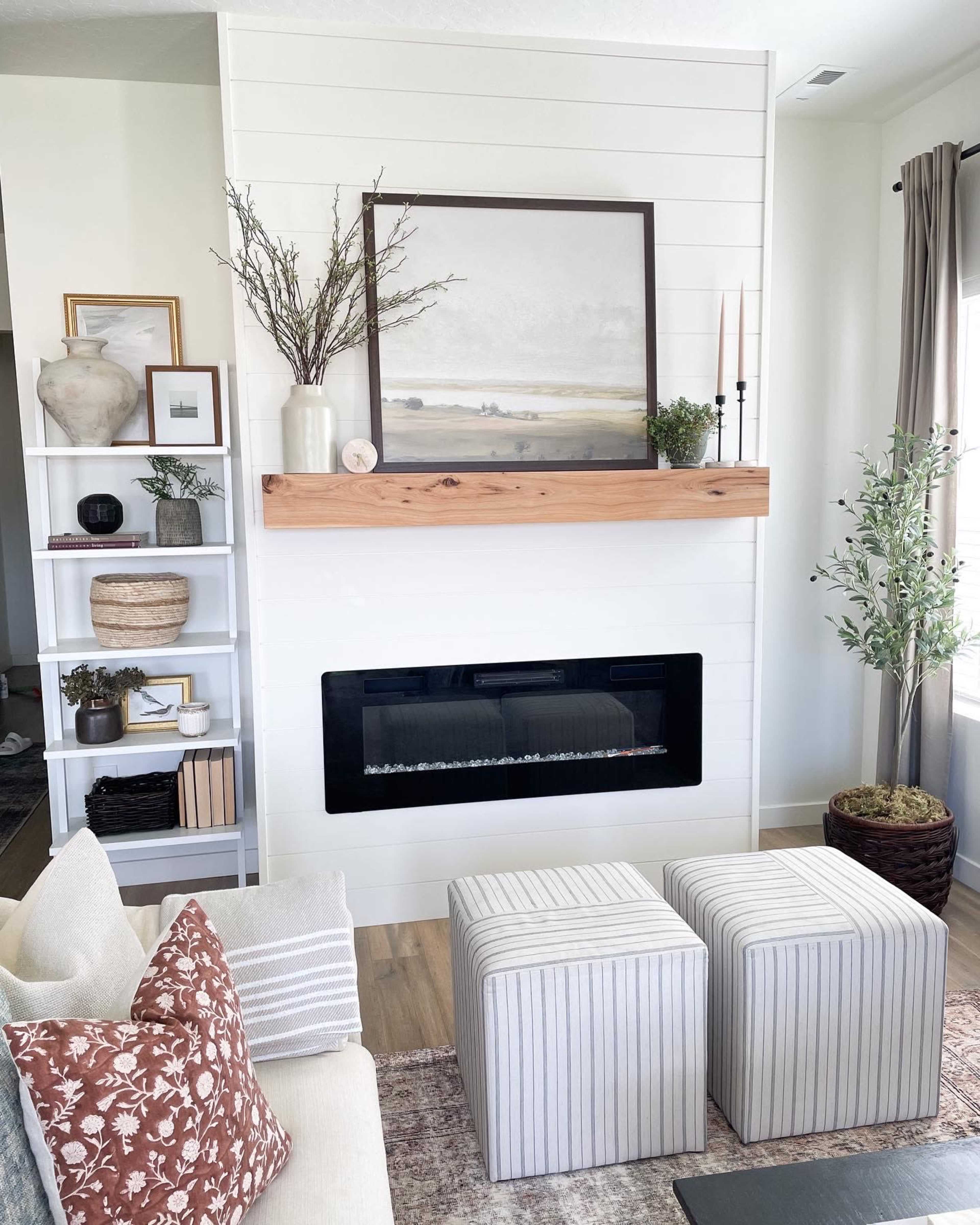 A modern living room with a fireplace built into a white shiplap wall, a wooden mantel, two striped ottomans, and decorative items on shelves nearby.