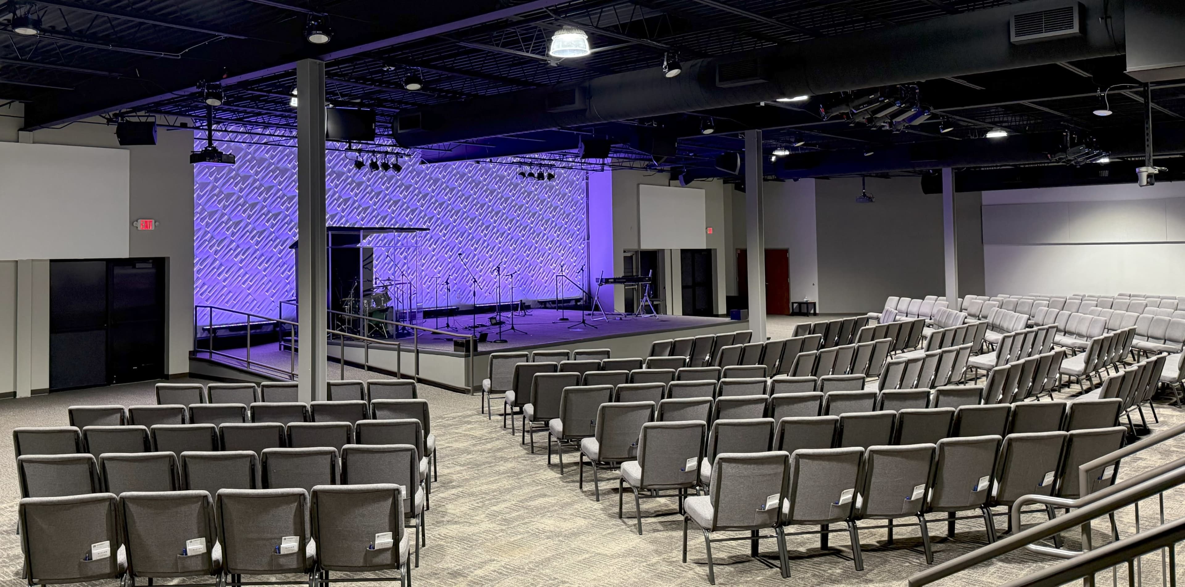 The image shows an interior view of a modern auditorium with rows of gray chairs facing a stage equipped with musical instruments.