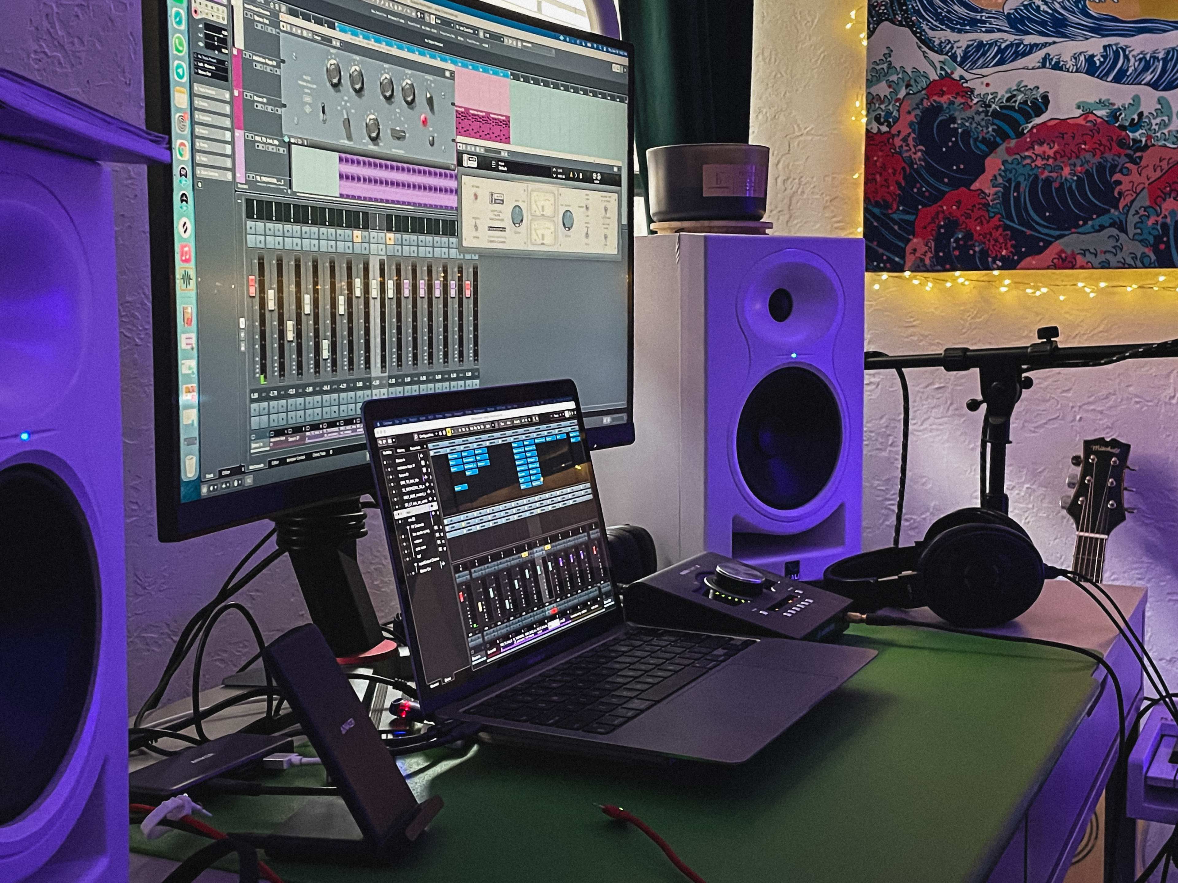 A computer setup for music production features a large monitor displaying audio software, a laptop, studio monitors, headphones, and a guitar on a desk.
