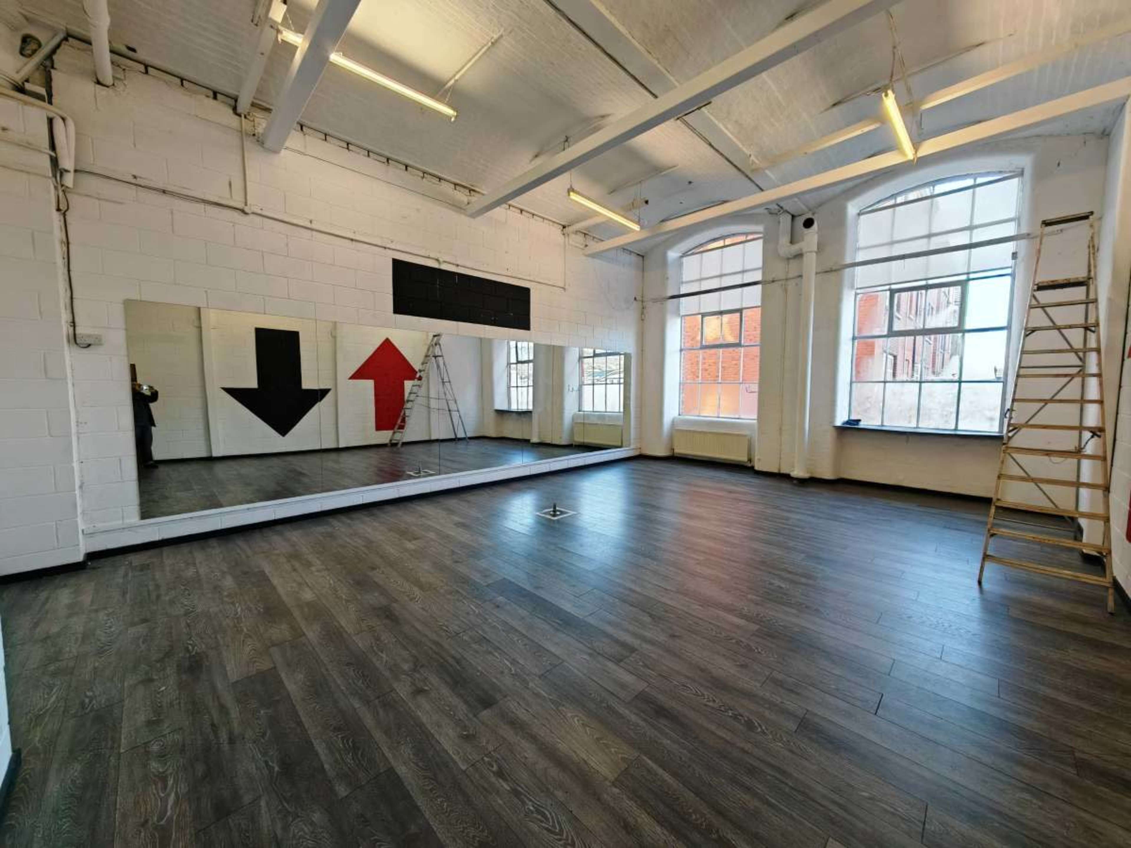 The image shows a spacious room with wooden flooring, large windows, a wall mirror, and two arrows painted on the wall, one black and one red.