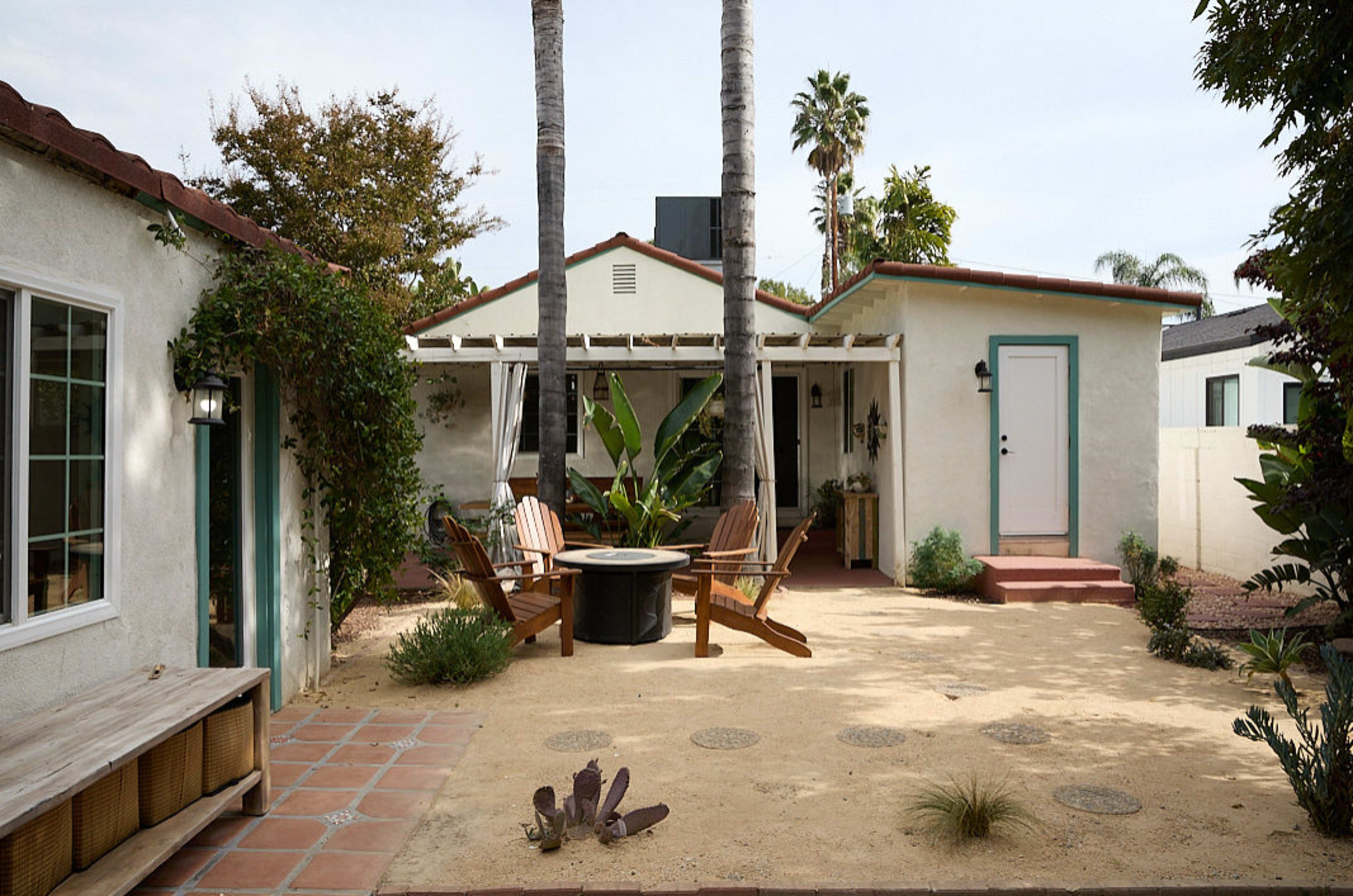 The image shows a quaint bungalow surrounded by a garden, featuring a circular seating area with wooden chairs and palm trees in the background.