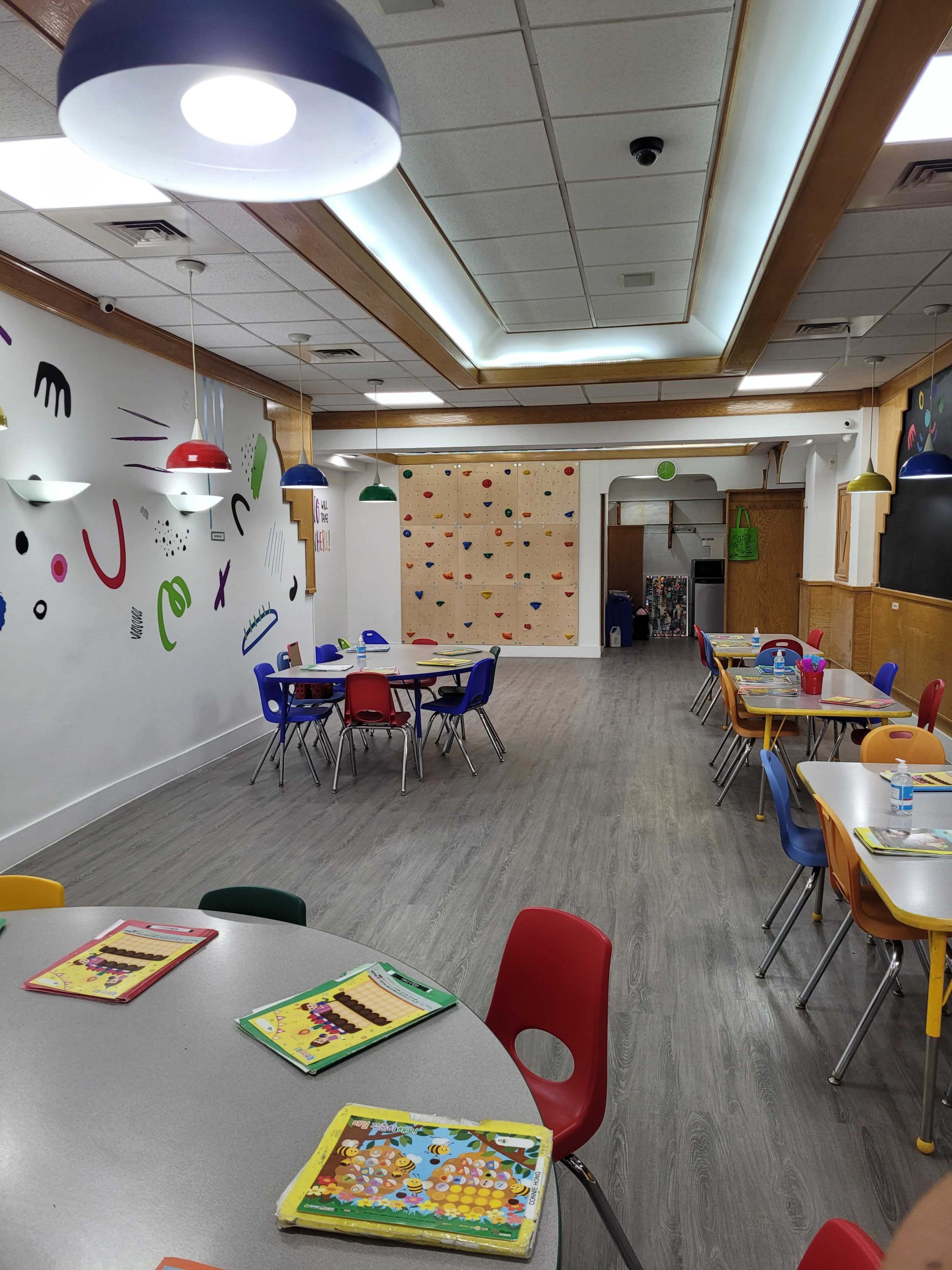 A brightly lit classroom features several round tables with colorful chairs, a decorated wall, and a section with bulletin boards.