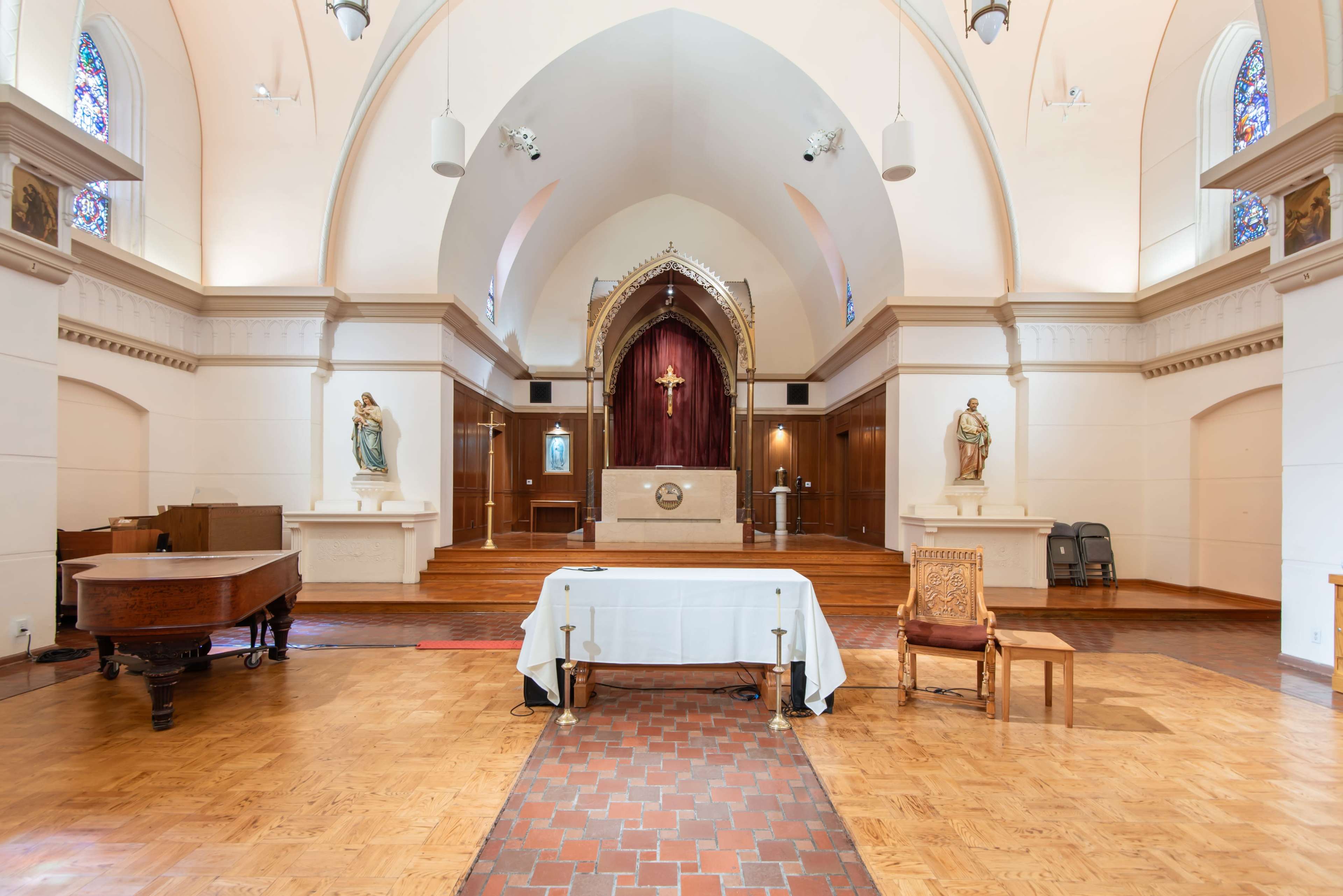 The image shows a church interior featuring a wooden altar with a red backdrop, flanked by two statues, and a grand piano in the foreground.