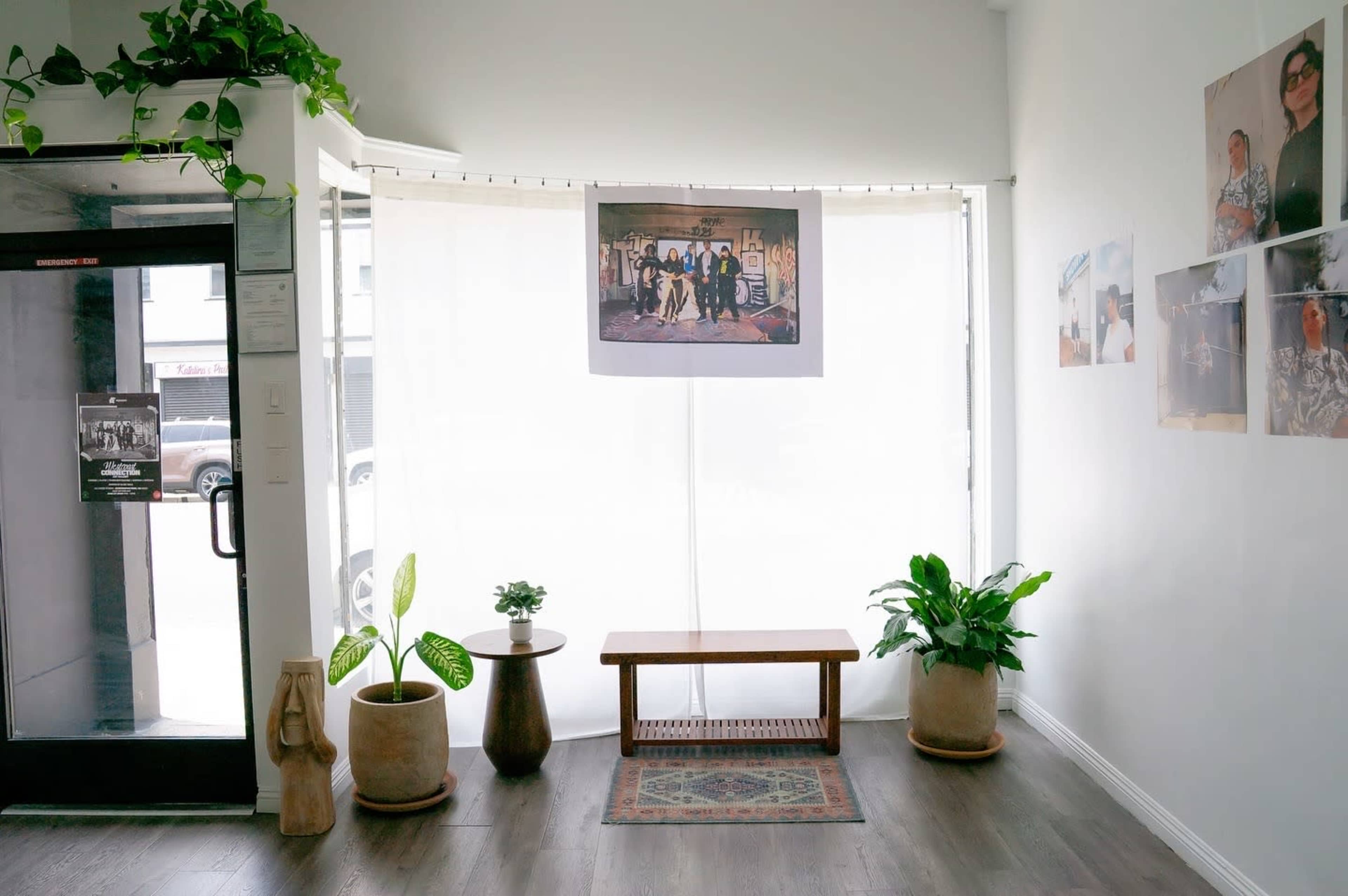 The interior of a bright room features a wooden bench, potted plants, a large picture hanging on a curtain, and a glass door leading outside.