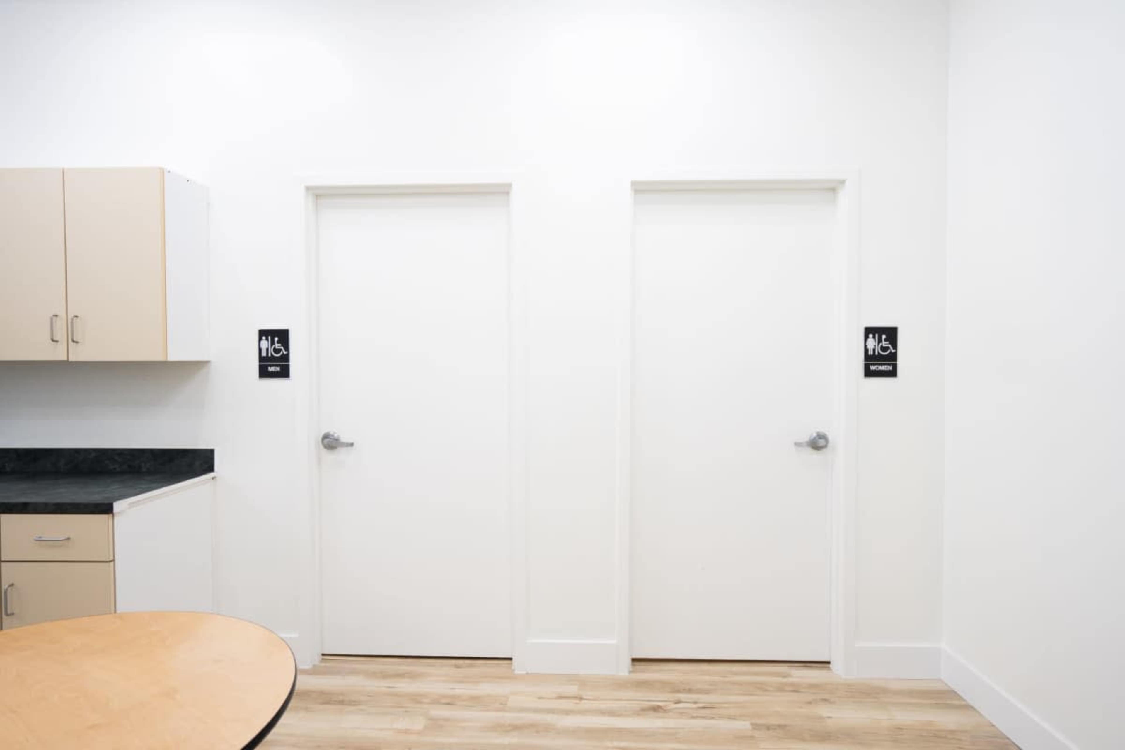 The image shows two closed white doors side by side, each labeled with signs indicating they are restrooms, in a bright room with a wooden floor and a kitchenette to the left.