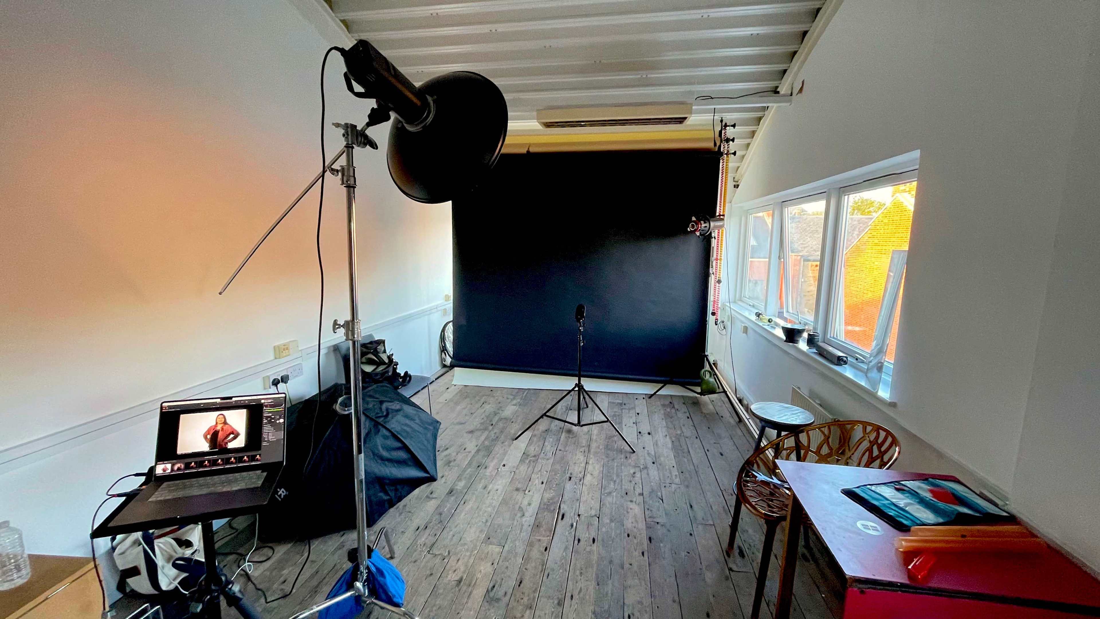 A photography studio with a black backdrop, lighting equipment, a microphone on a stand, and a laptop displaying a model.