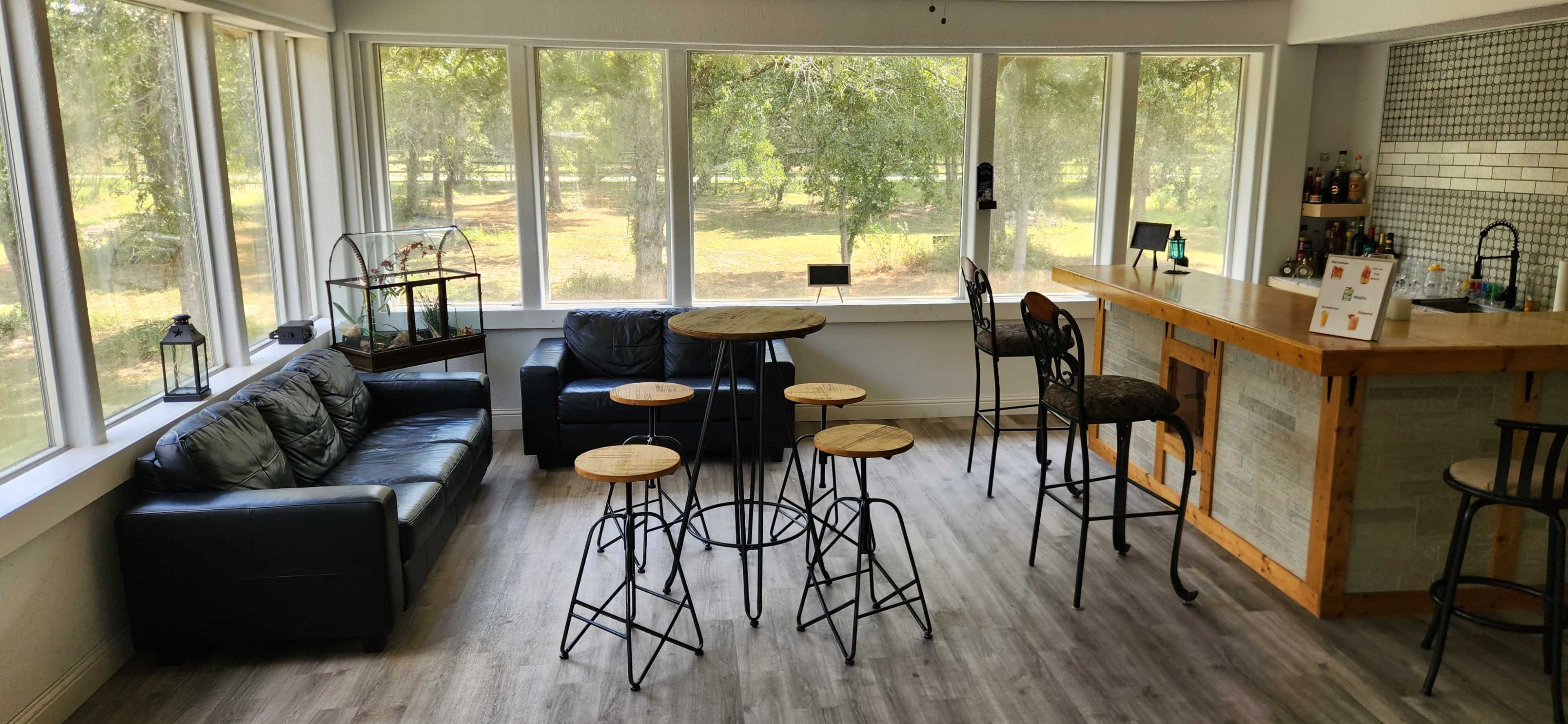 The image shows a sunlit lounge area with a leather couch, a round wooden table surrounded by four high stools, and a bar with a few bottles visible in the background.