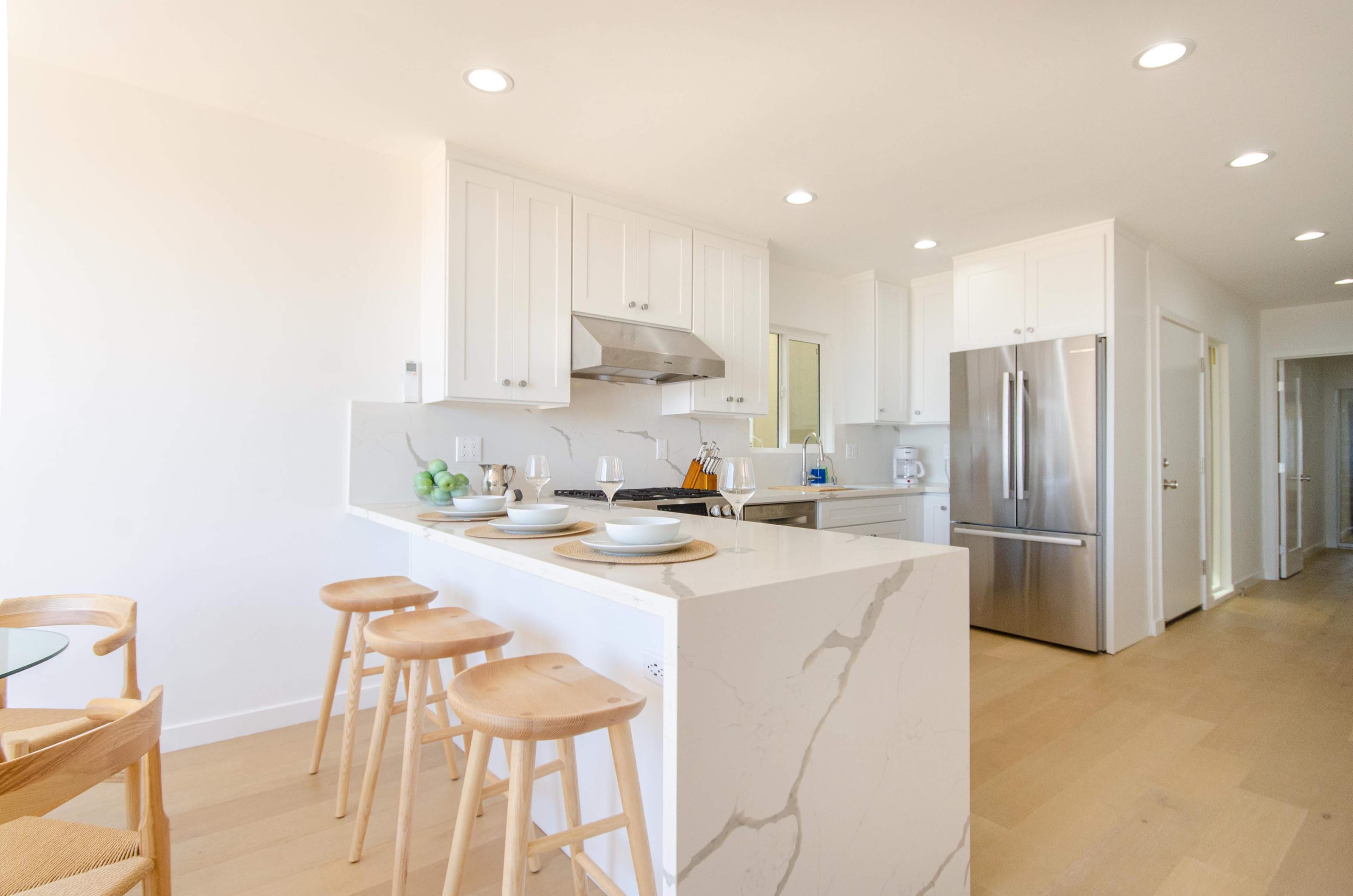A modern kitchen features white cabinetry, a large marble island with four wooden stools, and stainless steel appliances.