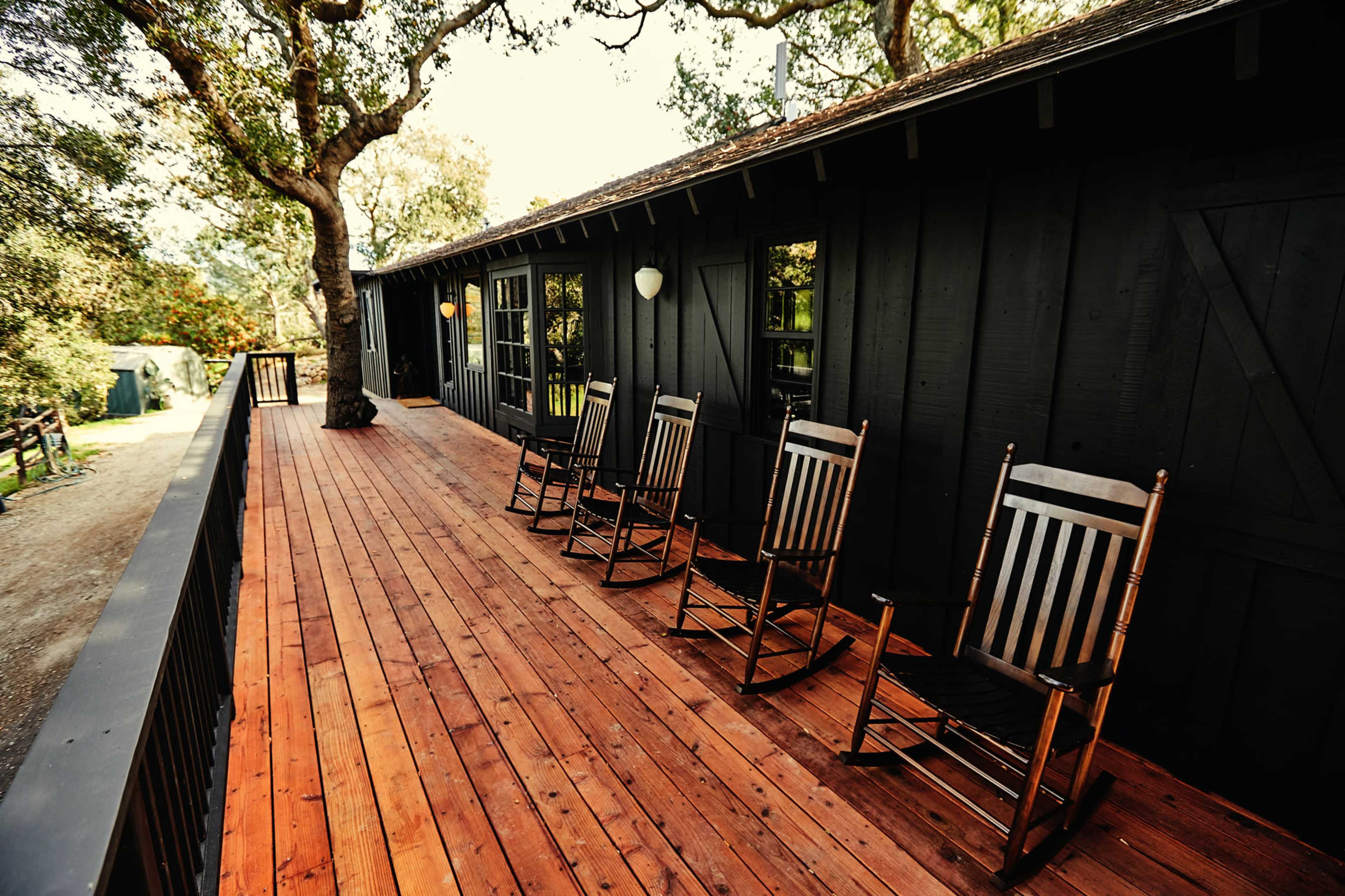 A row of wooden rocking chairs is positioned on a long, wooden deck attached to a dark-colored building.