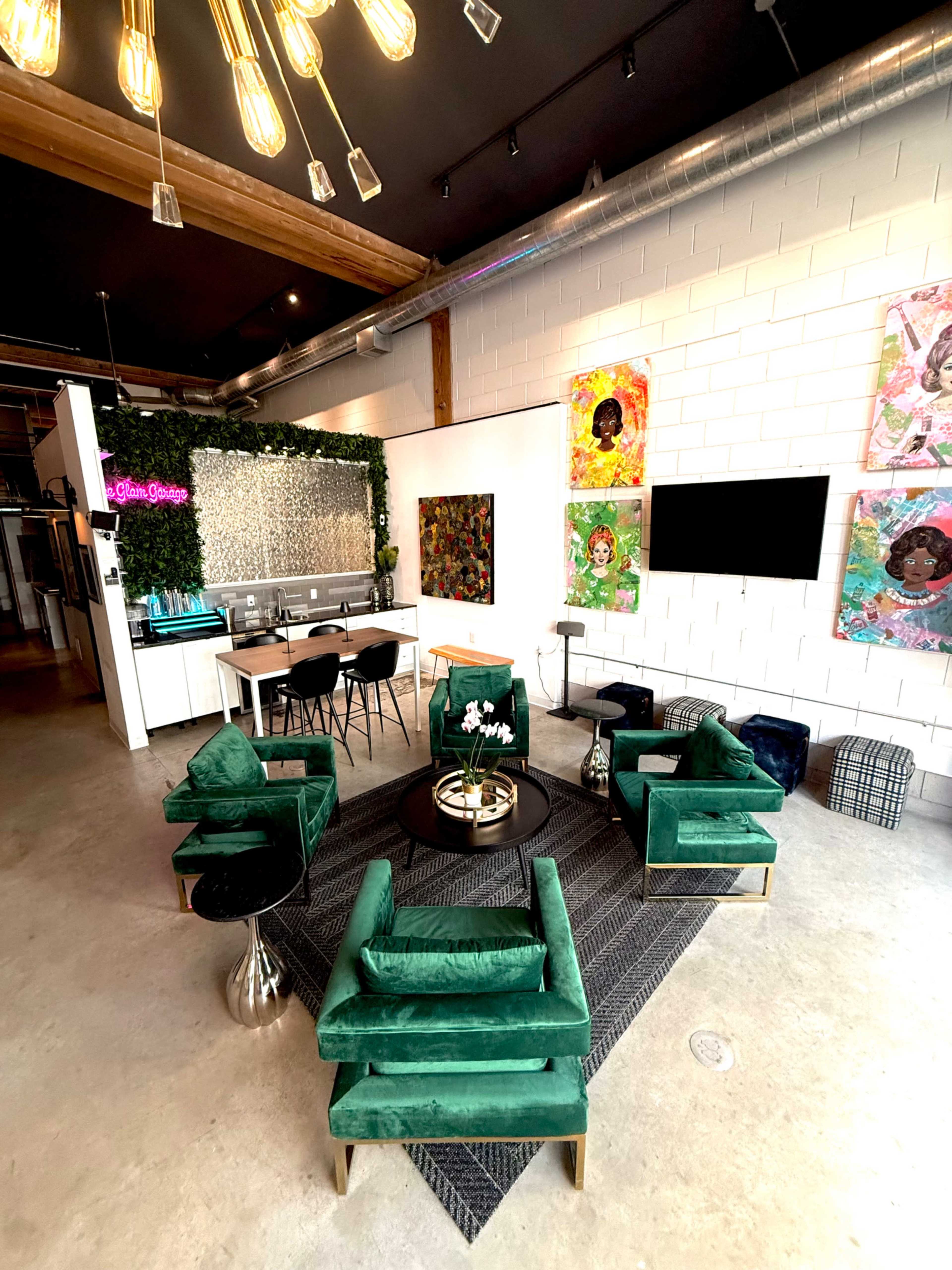 The image shows a modern lounge area with green velvet chairs arranged around a circular coffee table, a black rug, and colorful artwork on the walls.