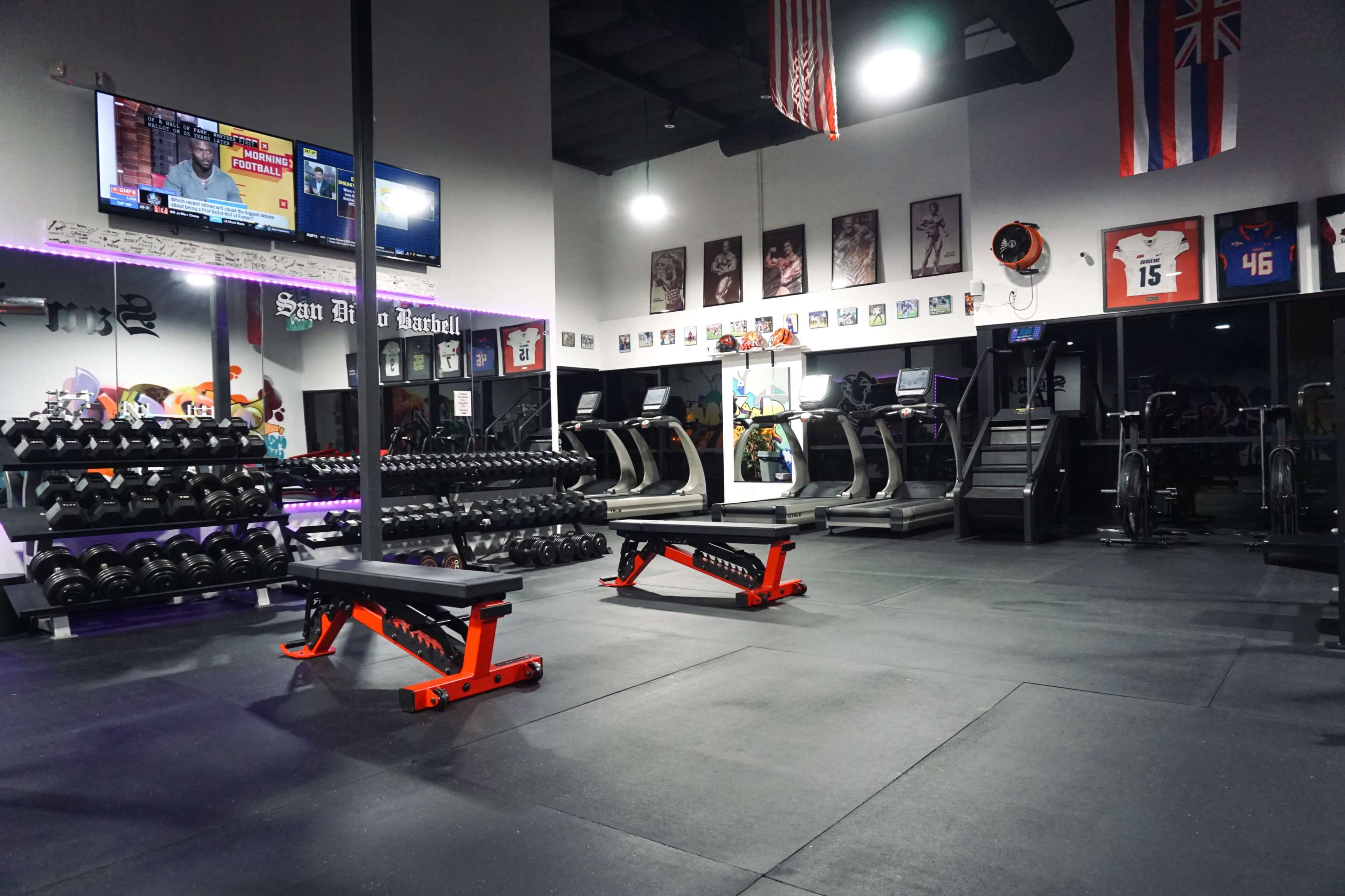 The image shows the interior of a gym featuring weights, benches, treadmills, and wall decorations including sports banners and framed photos.