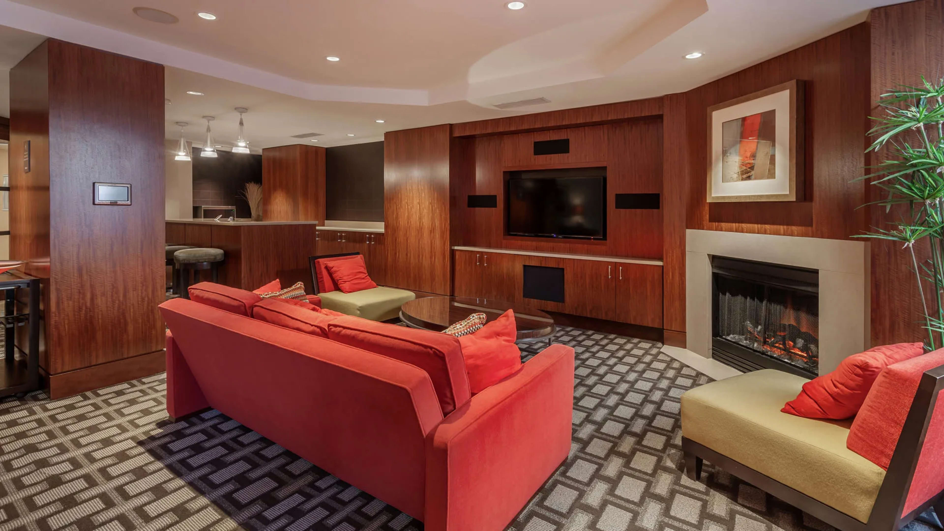 A modern living room features a red sofa with cushions, an area rug, a fireplace, and a wall-mounted television, surrounded by wooden paneling and a kitchenette in the background.
