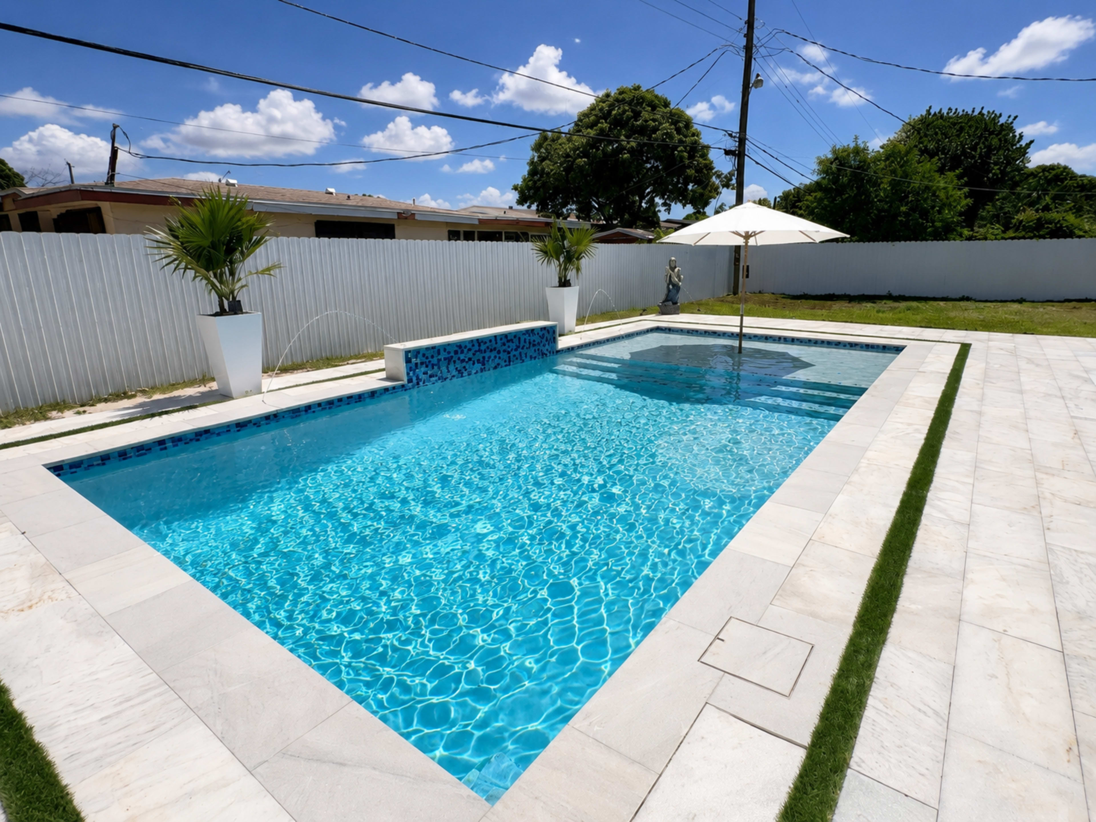 A rectangular swimming pool with clear blue water is situated in a well-maintained yard, surrounded by white fencing and dotted with palm trees and an umbrella.