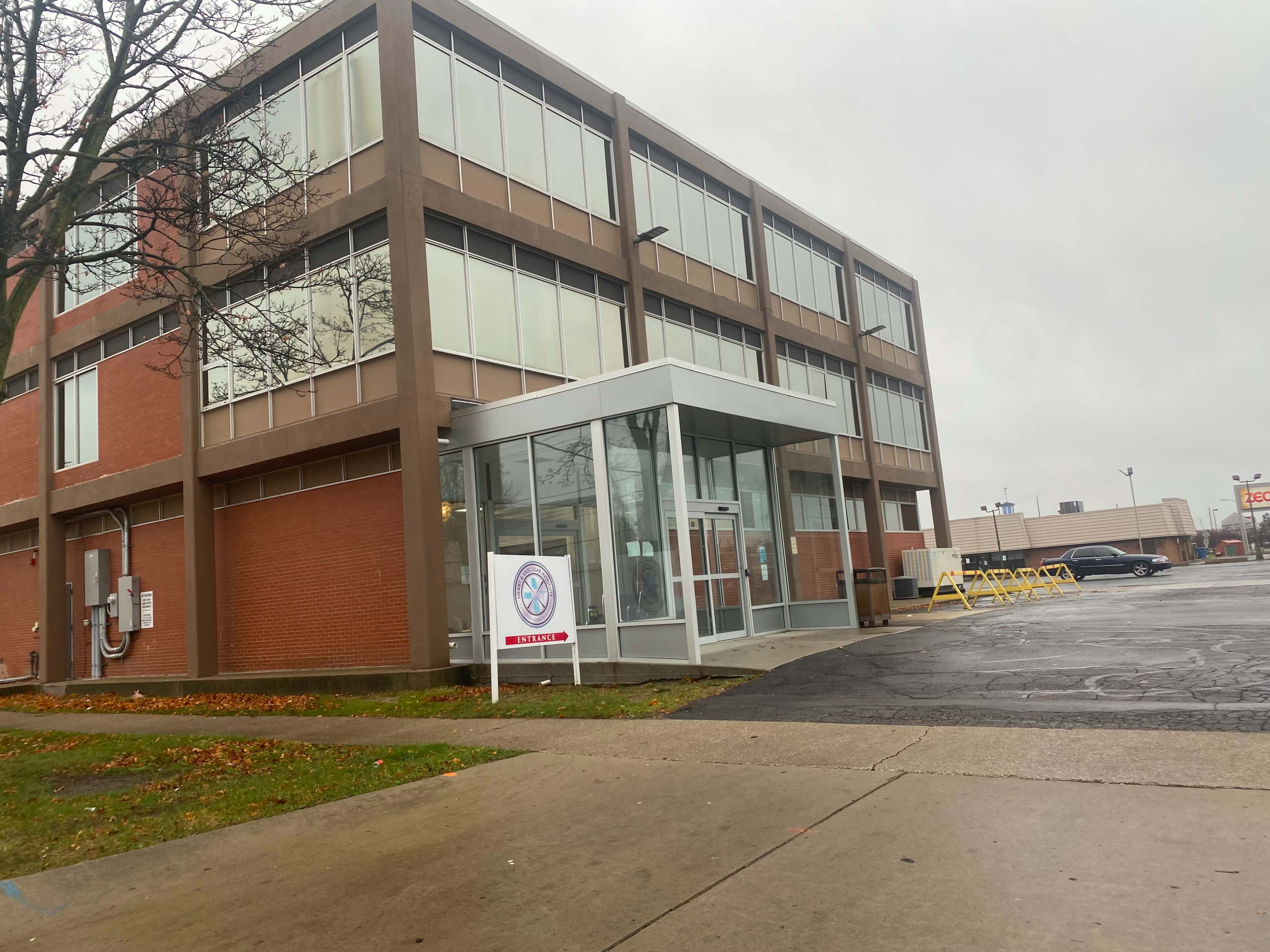 A modern, three-story building with large glass windows is located at a street corner, featuring a covered entrance and a parking lot.