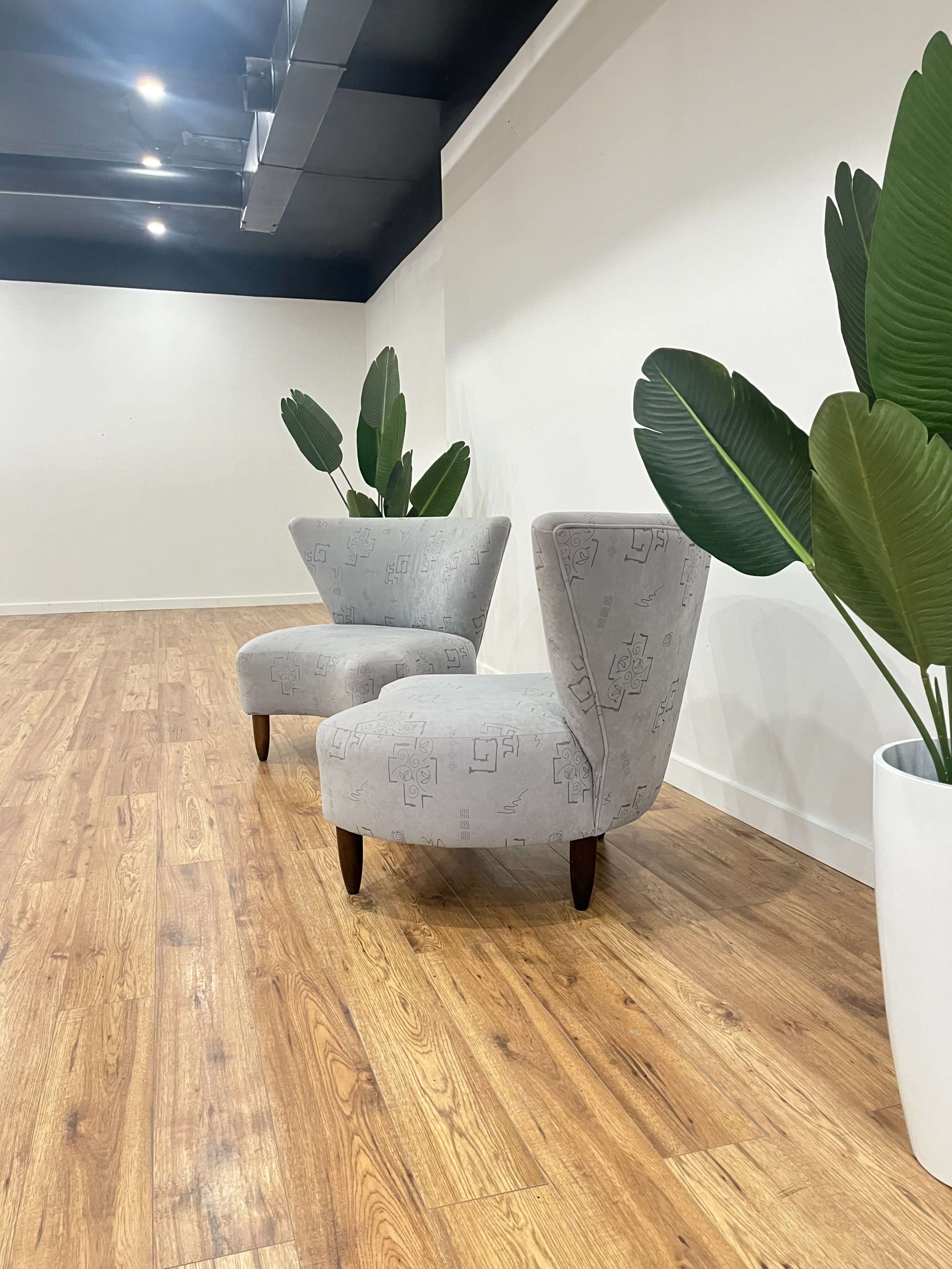 Two gray upholstered chairs with curved backs are positioned next to large potted plants in a modern indoor space with wooden flooring.