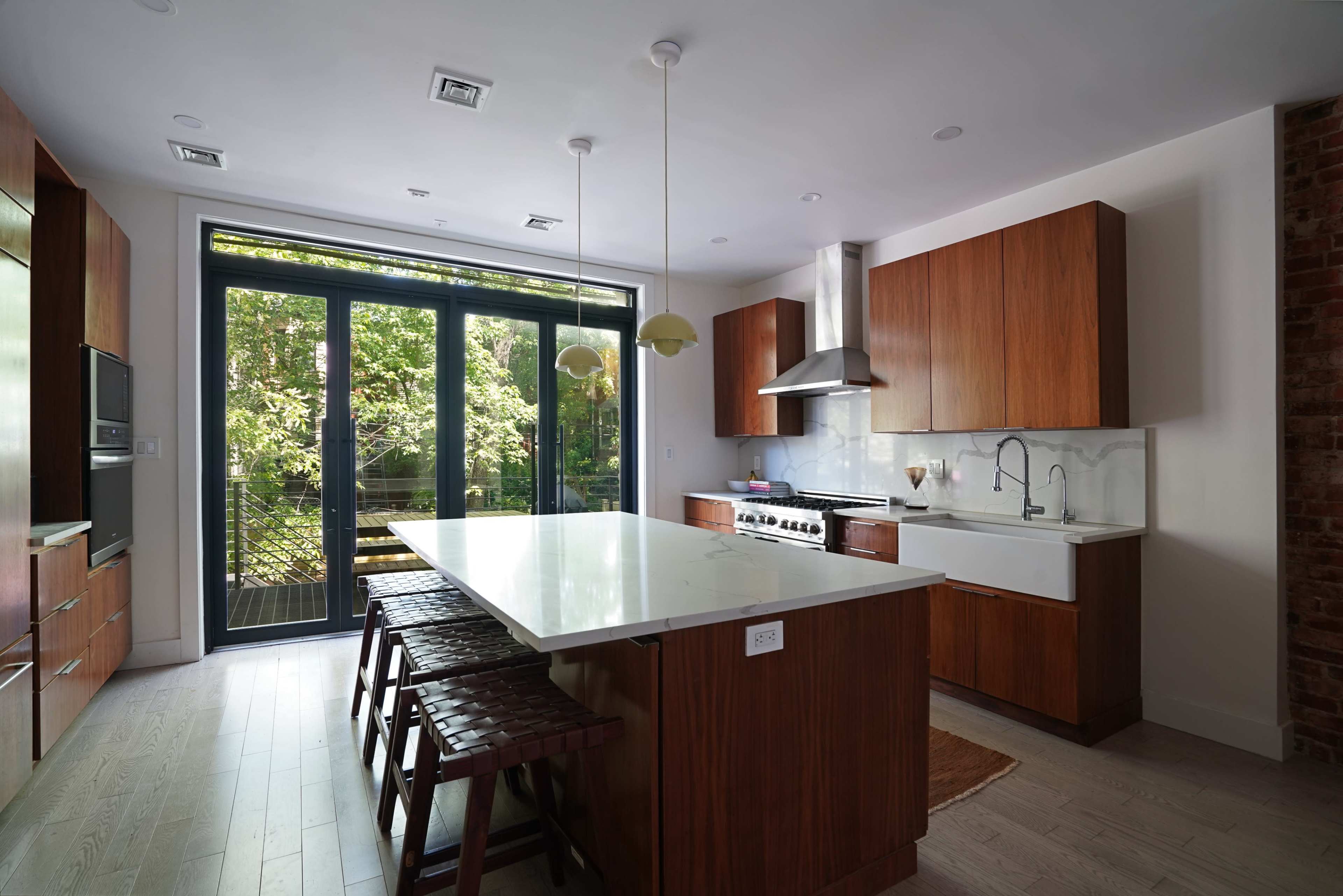 A modern kitchen features a large island with bar stools, wooden cabinetry, stainless steel appliances, and large windows overlooking a green outdoor space.