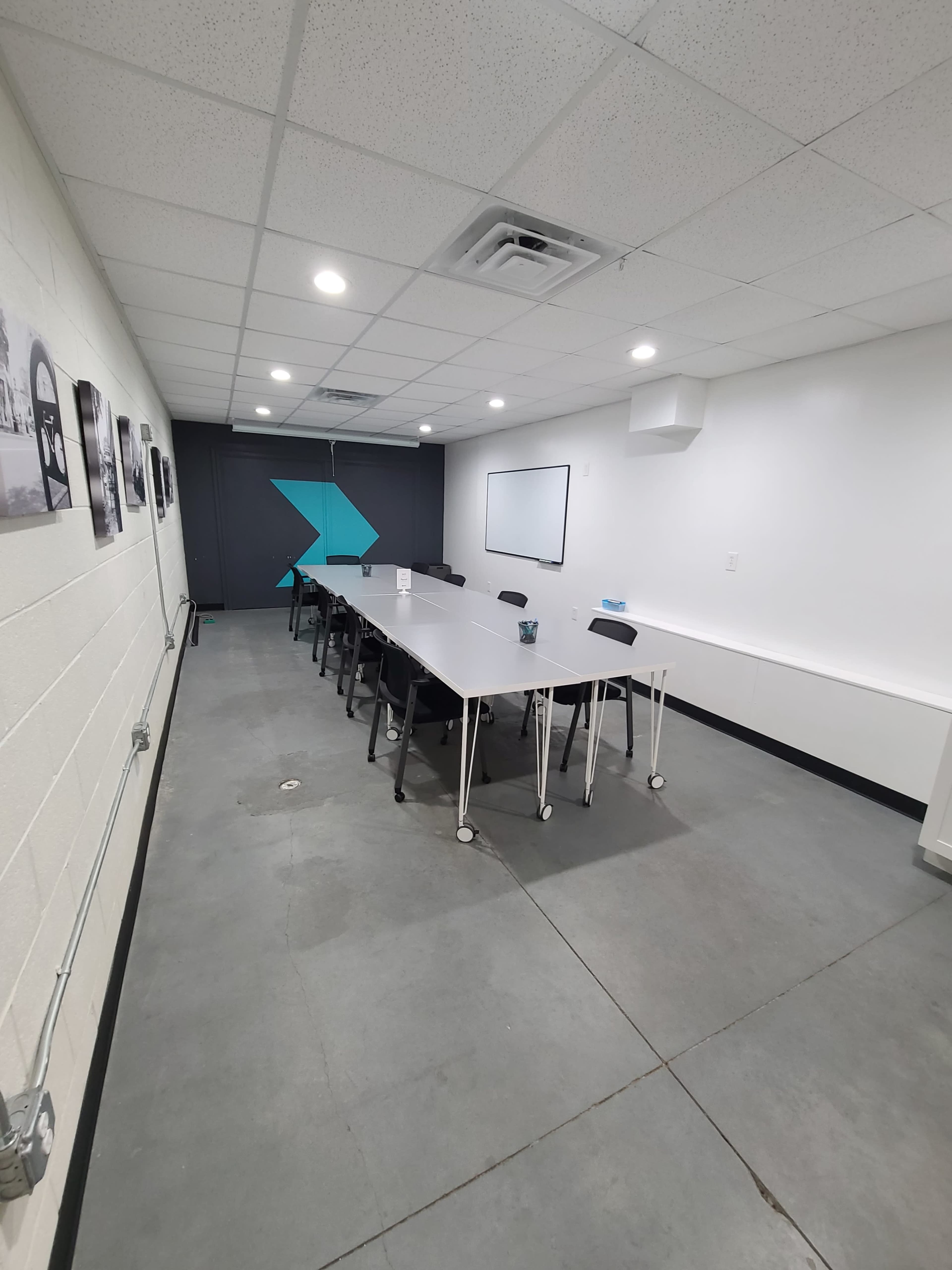 A long, rectangular conference table with chairs is positioned in a modern, minimalist meeting room featuring grey concrete floors and a wall with a large blue graphic.