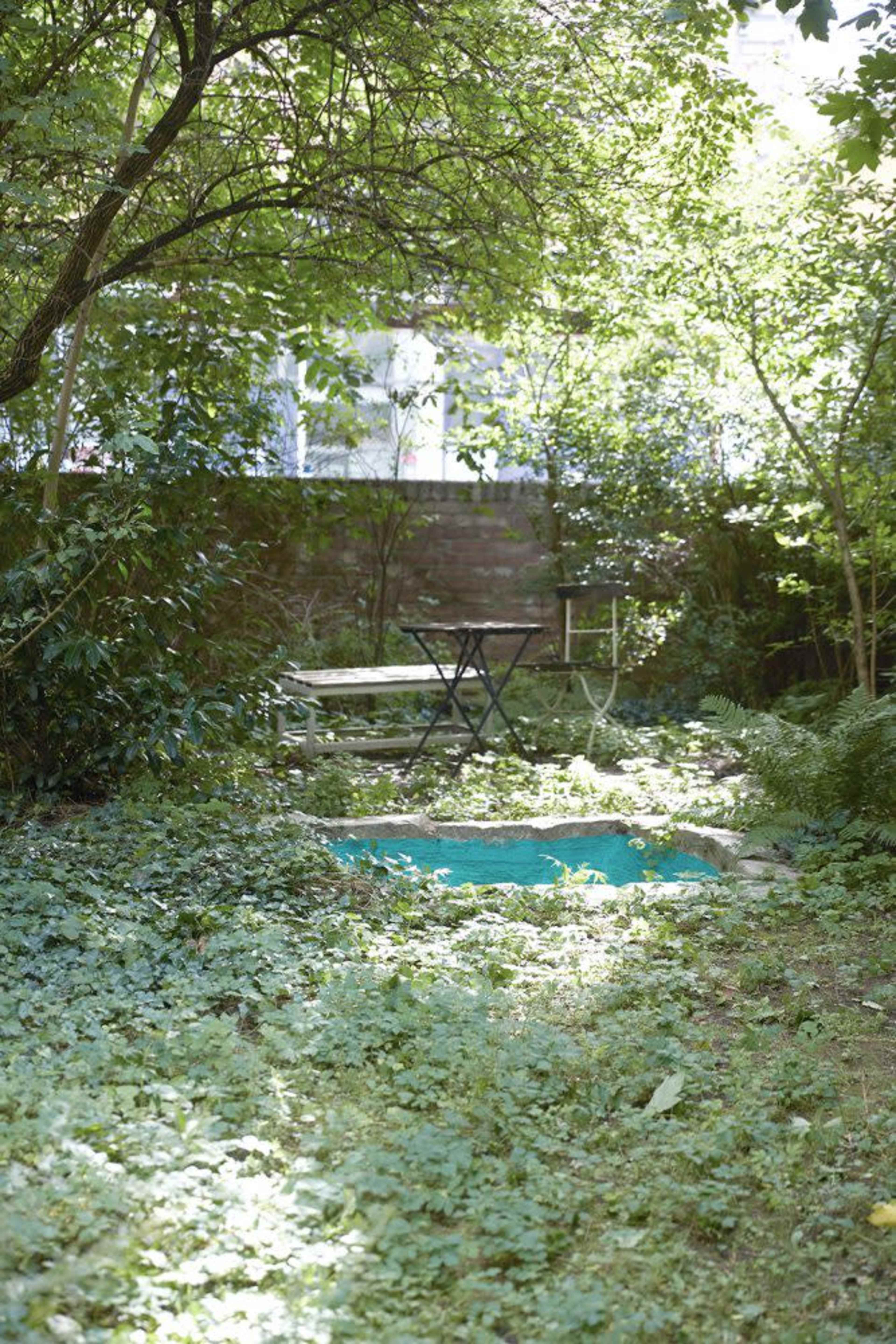 A small, secluded garden area features a simple table and chairs under a canopy of trees, surrounded by lush greenery.