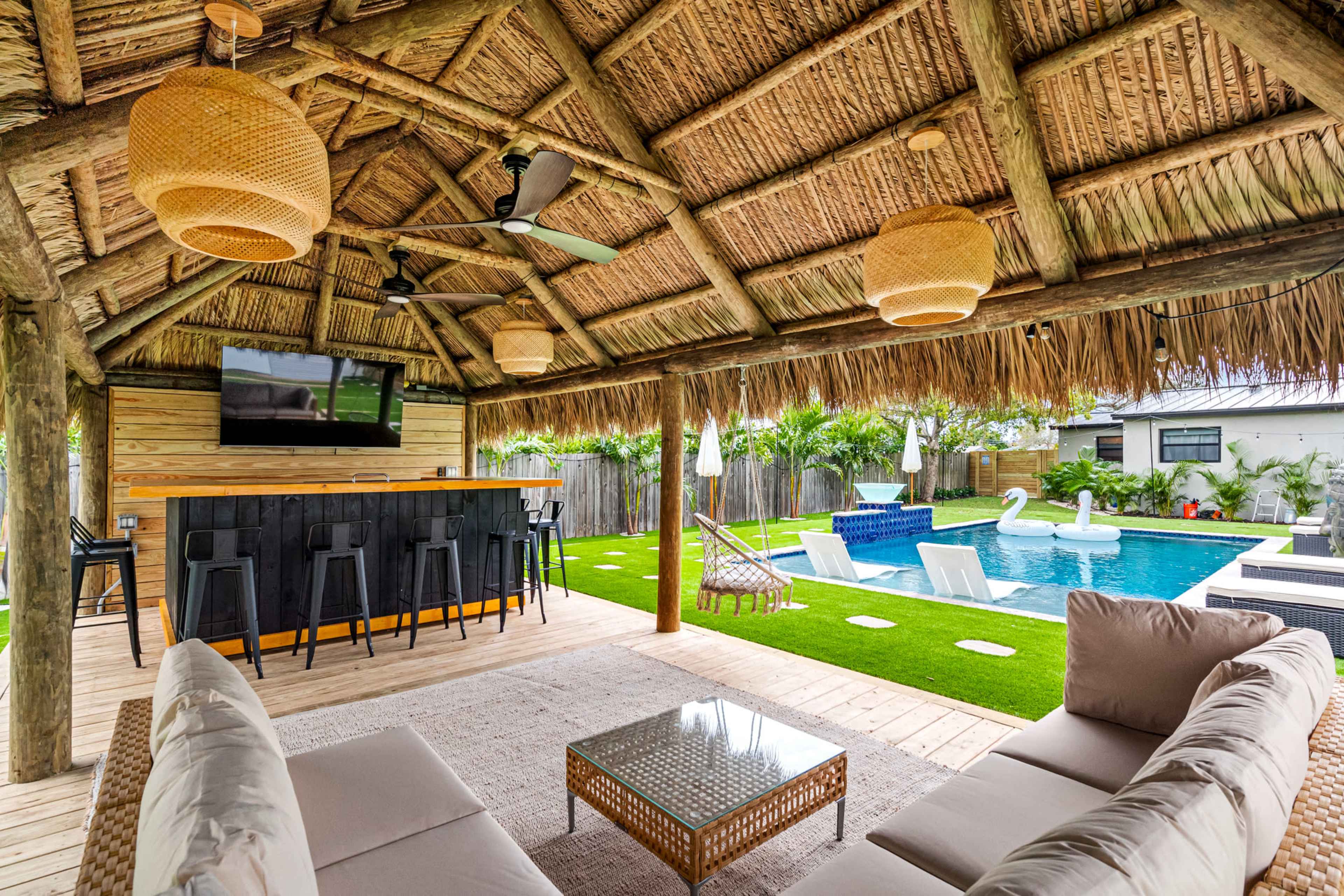 A thatched-roof patio features a lounge area with a couch and coffee table, overlooking a swimming pool equipped with inflatable swans and a bar area with seating.