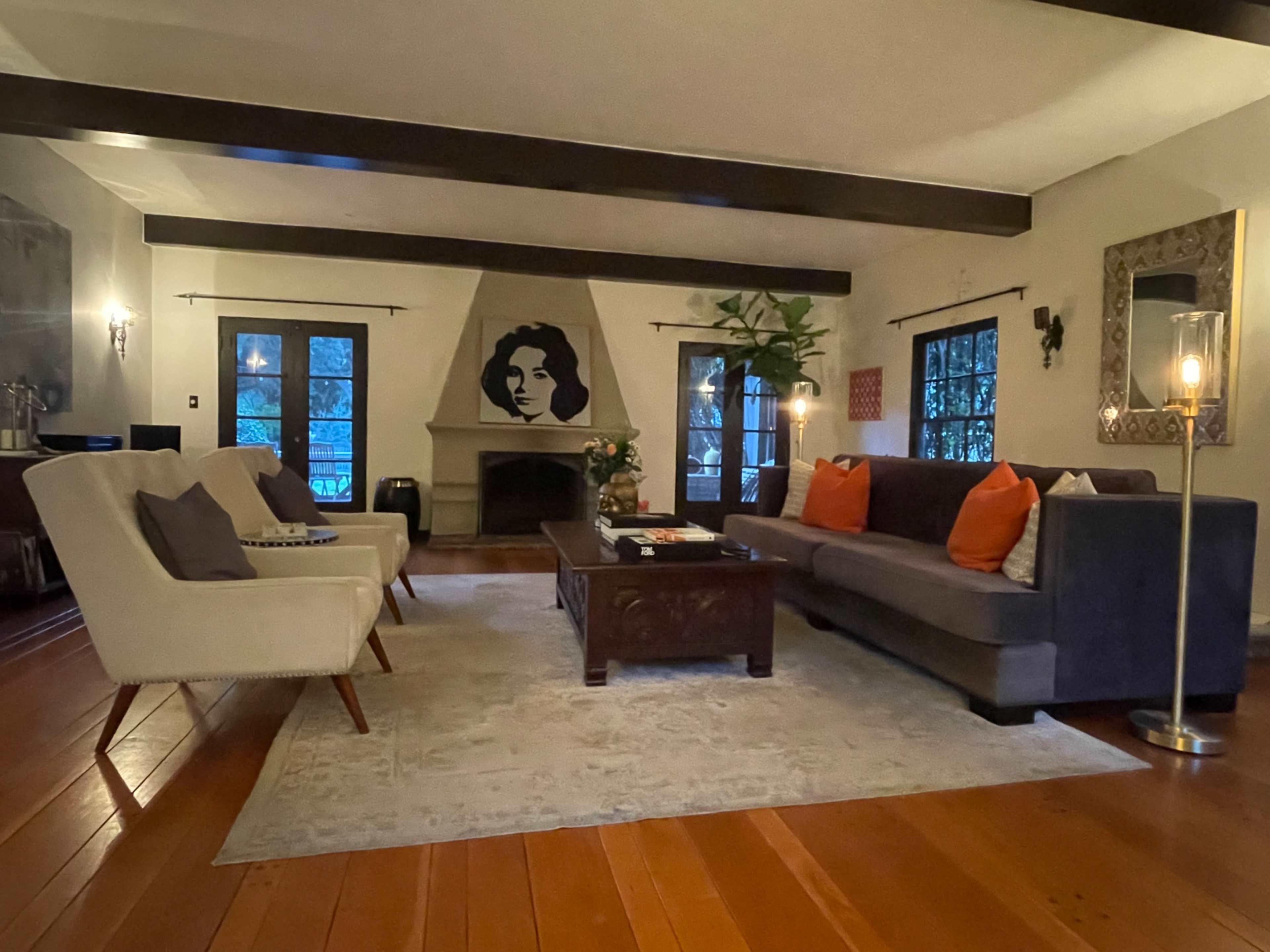 The living room features a dark sofa adorned with orange pillows, a decorative area rug on wooden flooring, a wall mural of a woman's face, and large windows framed by black curtains.