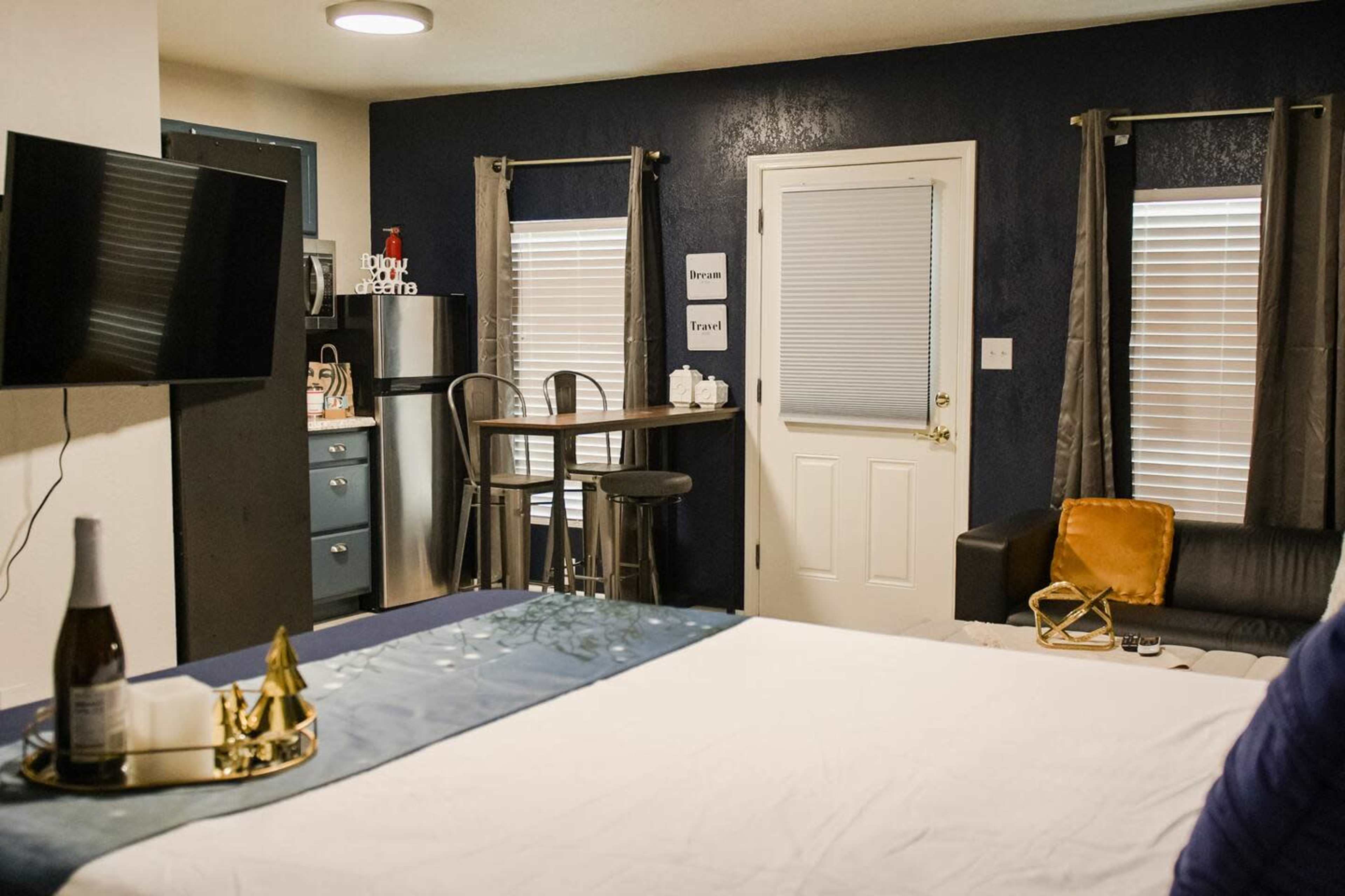 The image shows a small, modern studio room featuring a bed, a TV mounted on the wall, a kitchenette area with bar stools, and a door leading to the outside.