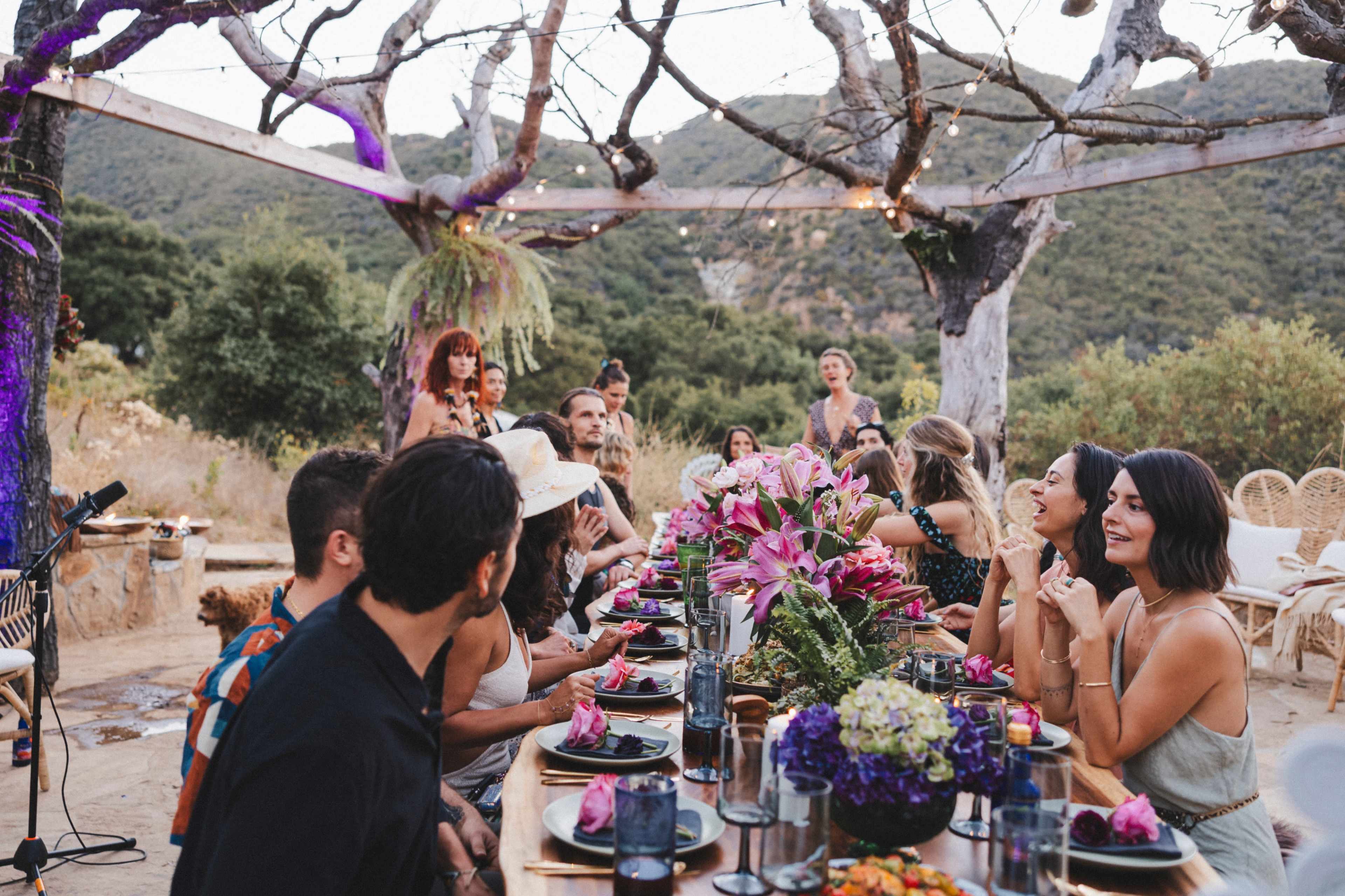 A large outdoor gathering features a long table set with food and flowers, surrounded by guests enjoying the evening ambiance beneath a rustic wooden structure and trees.