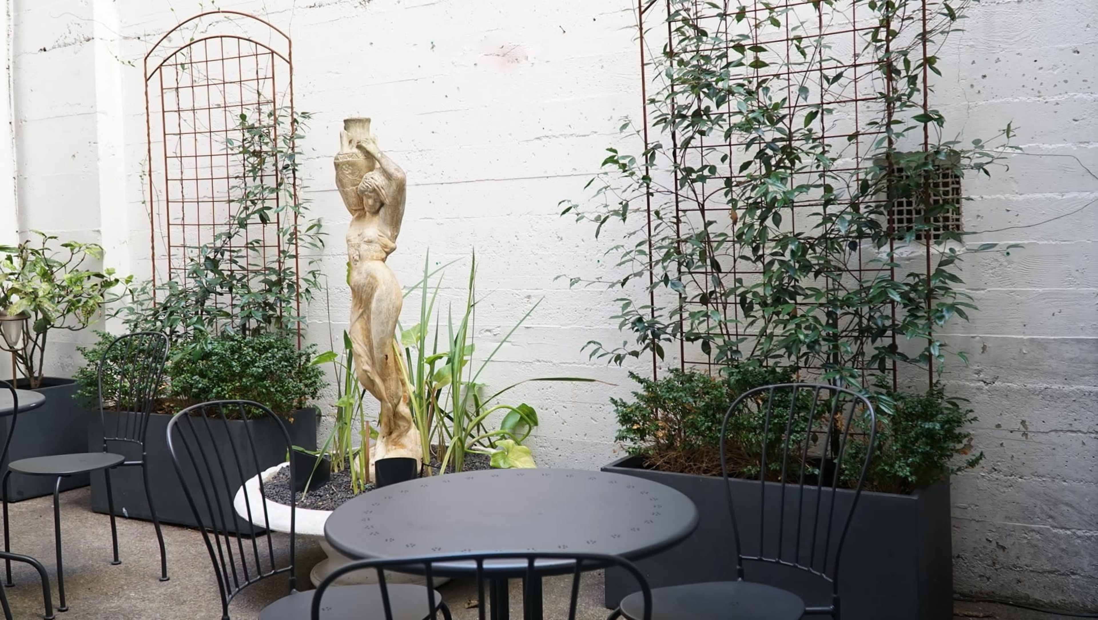 A courtyard features a round black table and chairs surrounded by greenery, including planters and trellises, with a sculpture of a figure in the center.