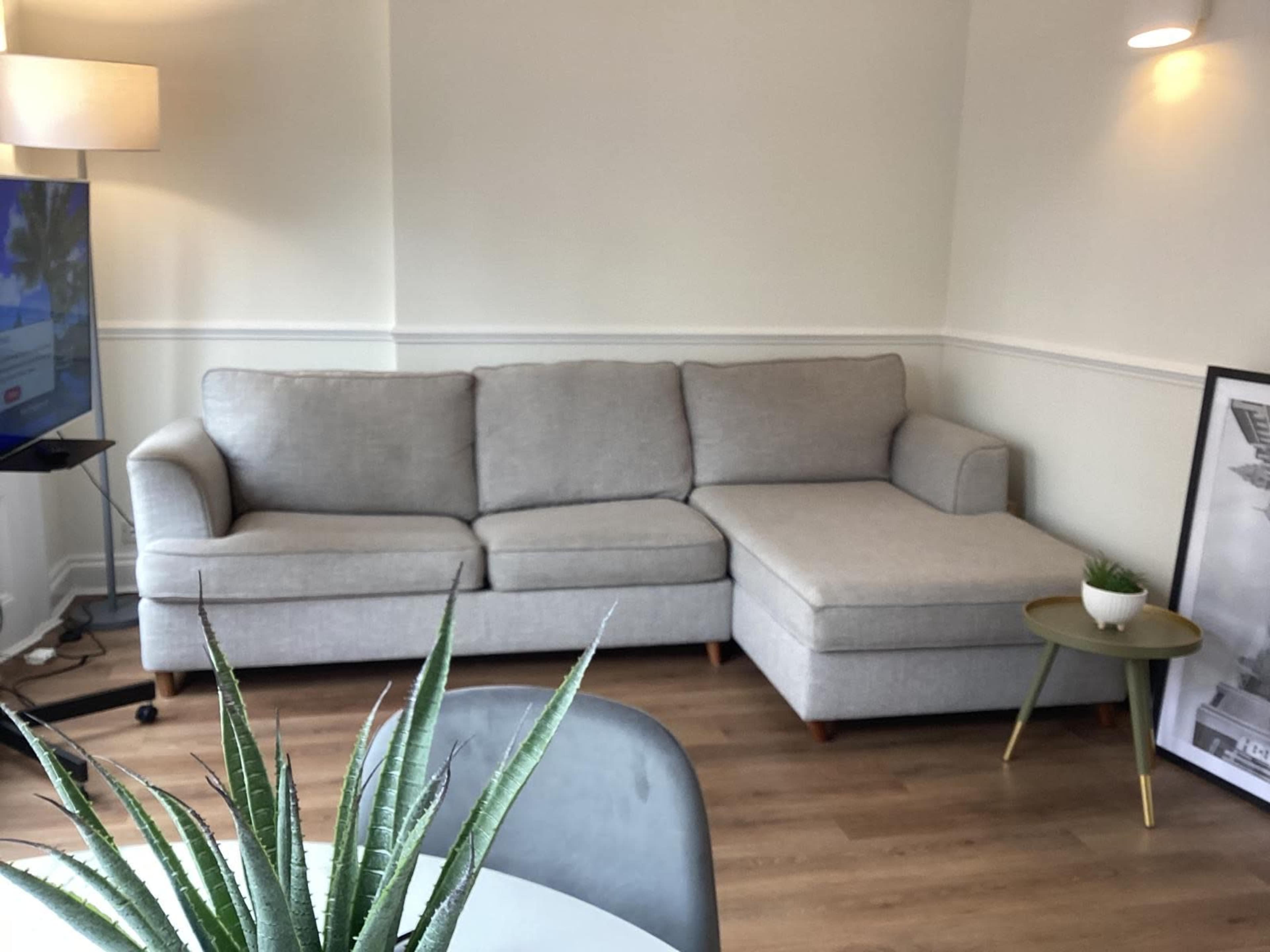 The image shows a light gray sectional sofa in a softly lit corner of a room with wooden flooring and a small table beside it.