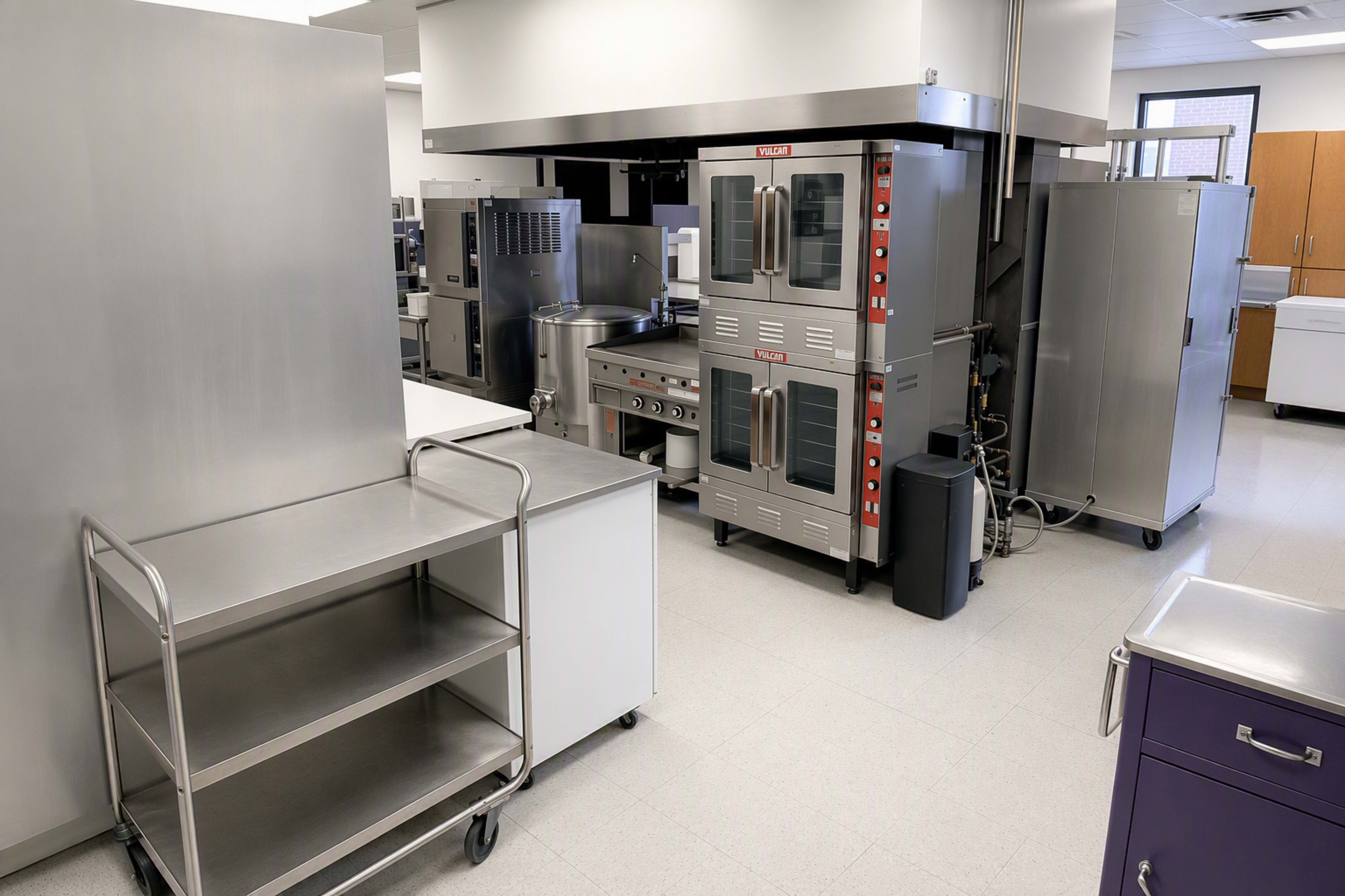 The image shows a commercial kitchen with stainless steel appliances, including ovens and a stove, along with a metal cart and a refrigerator.