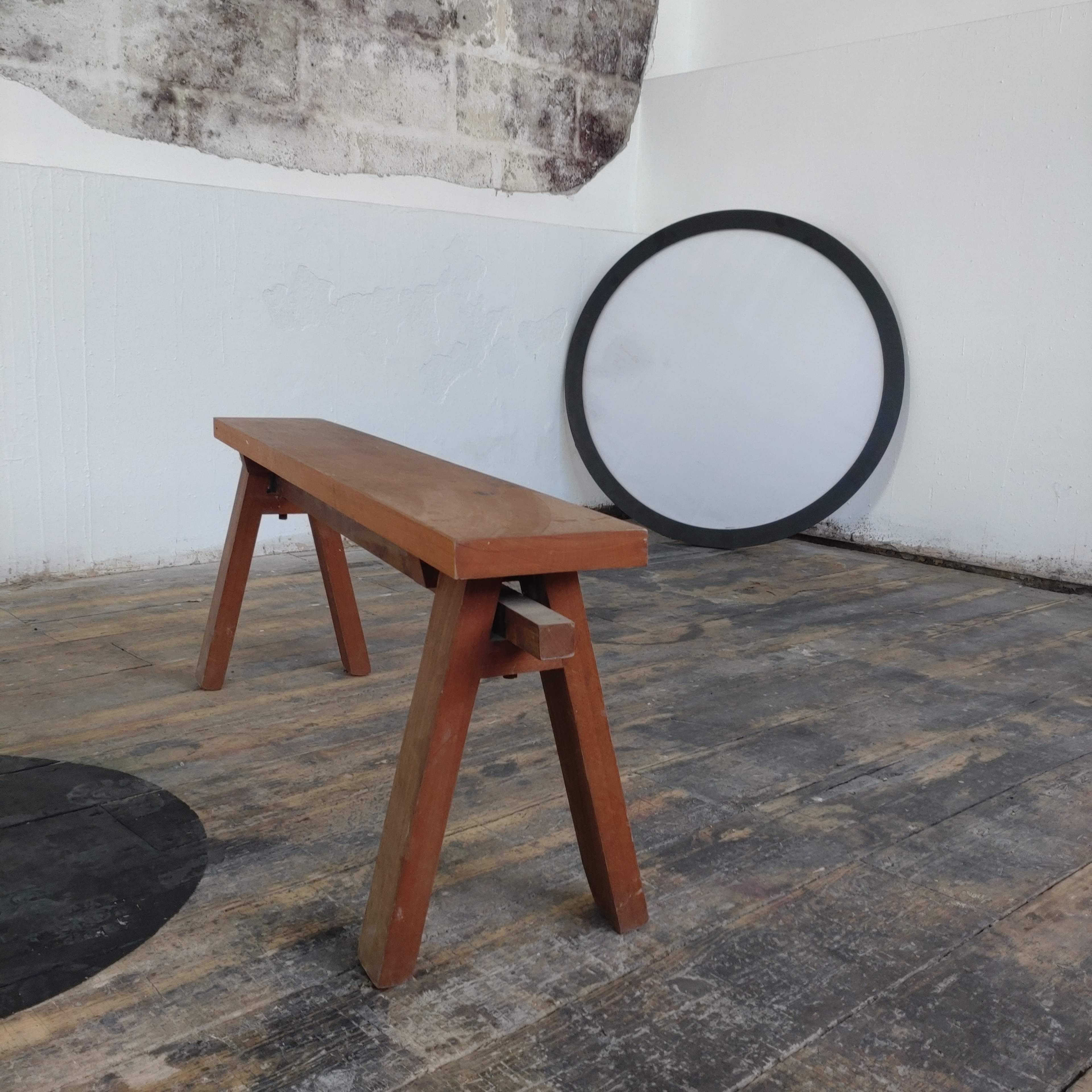 A wooden bench placed on a wooden floor, with a circular object and a textured wall in the background.