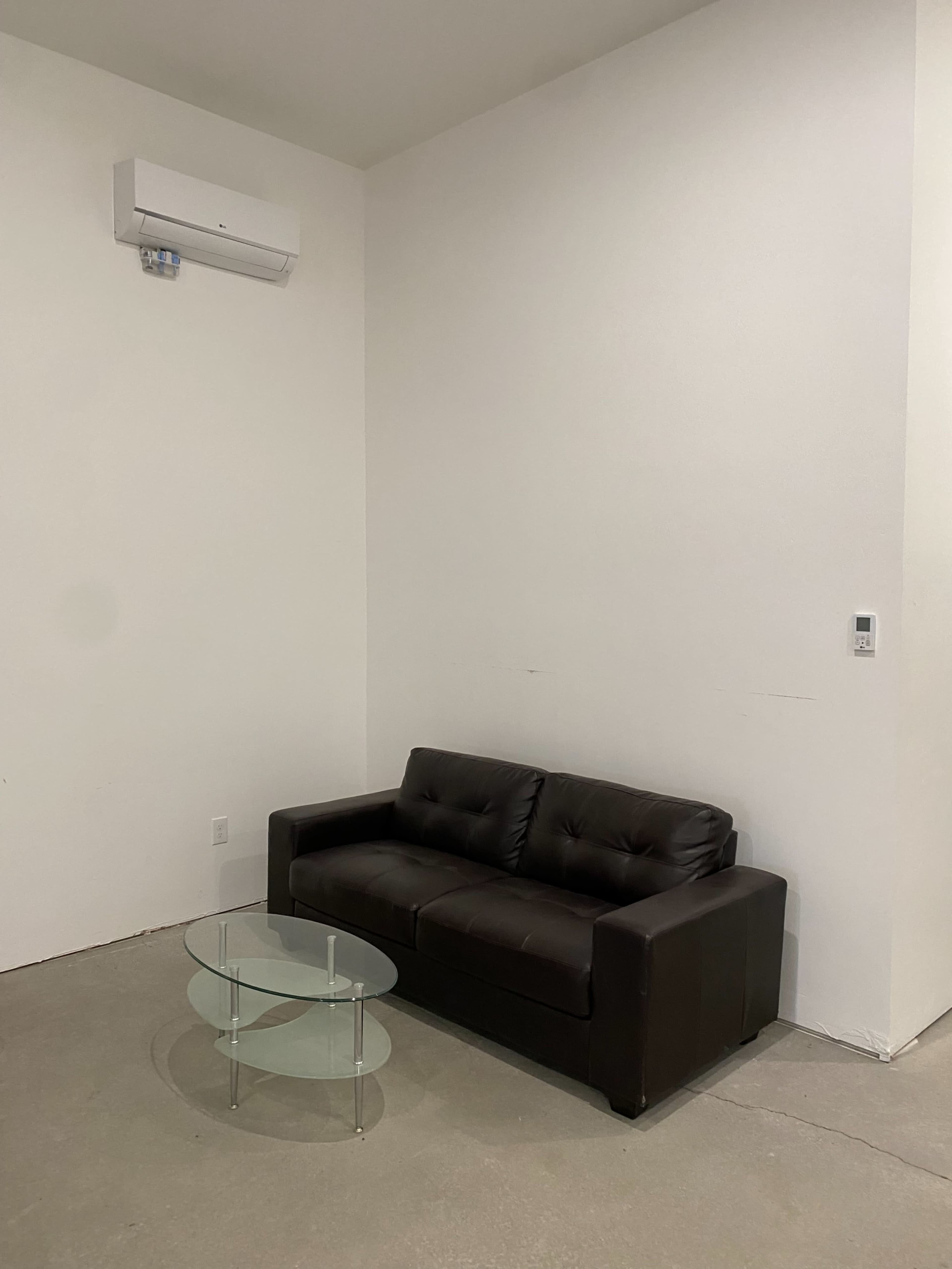 A black leather sofa is placed against a white wall next to a glass coffee table in a minimalist room with concrete flooring.