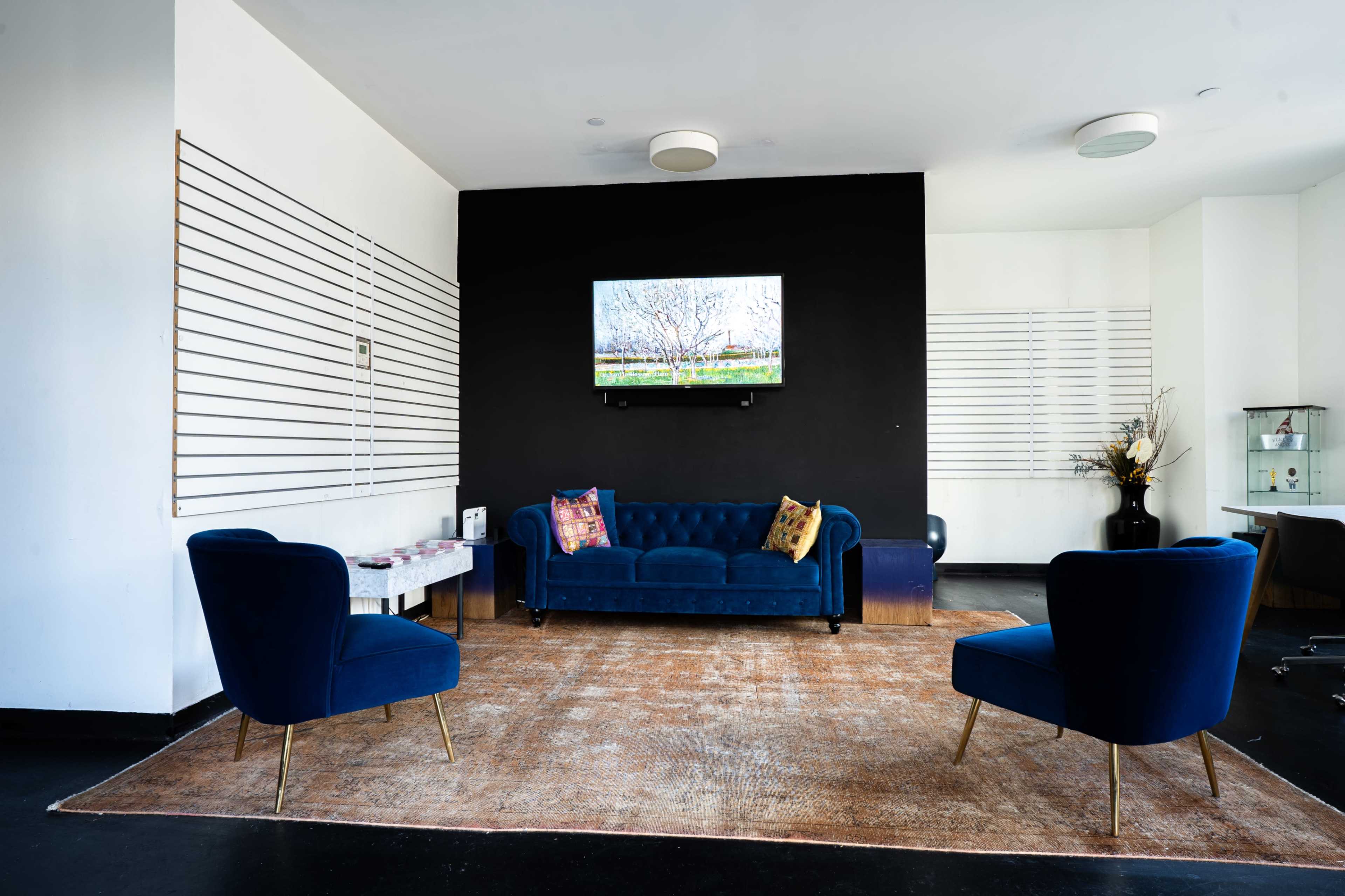 The image shows a modern living area with a navy blue sofa, two matching armchairs, a large area rug, and a wall-mounted television above a coffee table against a black accent wall.